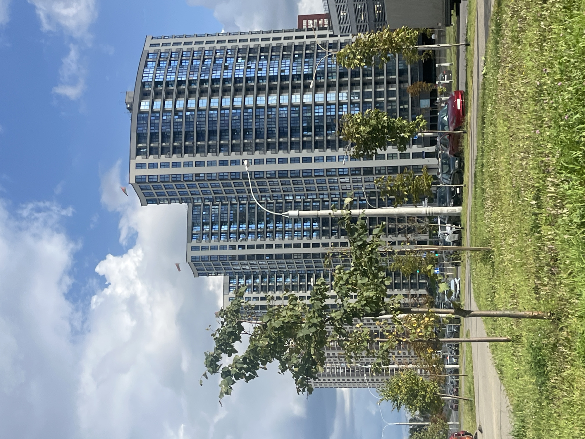 The image depicts a modern residential complex in Minsk, Belarus, characterized by tall, multi-story buildings with blue-tinted windows and balconies. The buildings are arranged in a row, suggesting a new development. In the foreground, there is a street with parked cars and a patch of grass with young trees. The sky is partly cloudy, with patches of blue sky visible. The overall impression is one of urban development and modern architecture in a city setting.