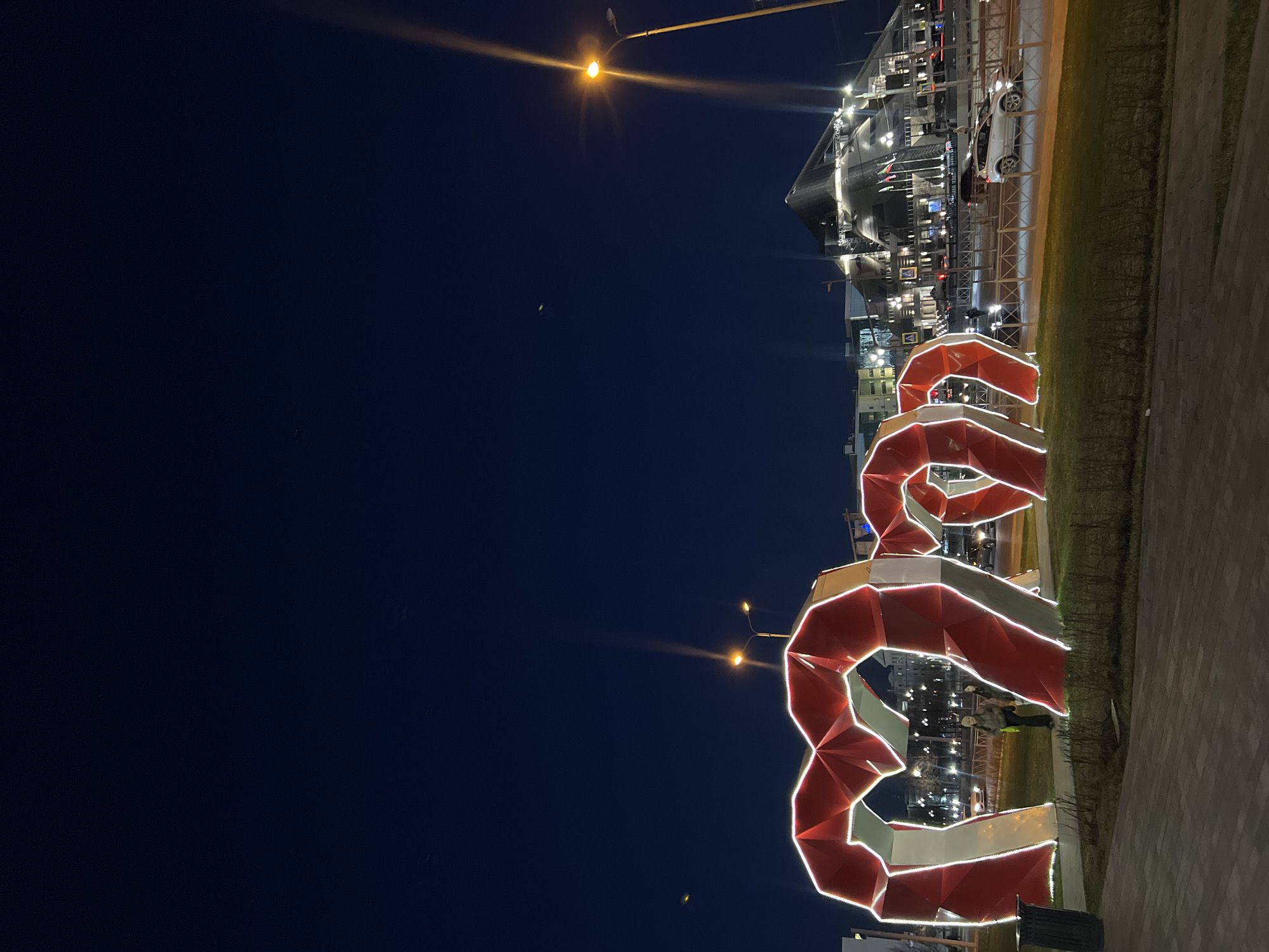 The image captures a nighttime scene in Kazan, Russia. A large, illuminated sculpture resembling a ribbon is prominently displayed on a paved area alongside a patch of grass. The sculpture is red and white with linear lighting. Behind the sculpture, a modern building with a dark glass facade is visible. Streetlights with a yellowish glow are also present. A white car is parked near the modern building. The background sky is dark blue.