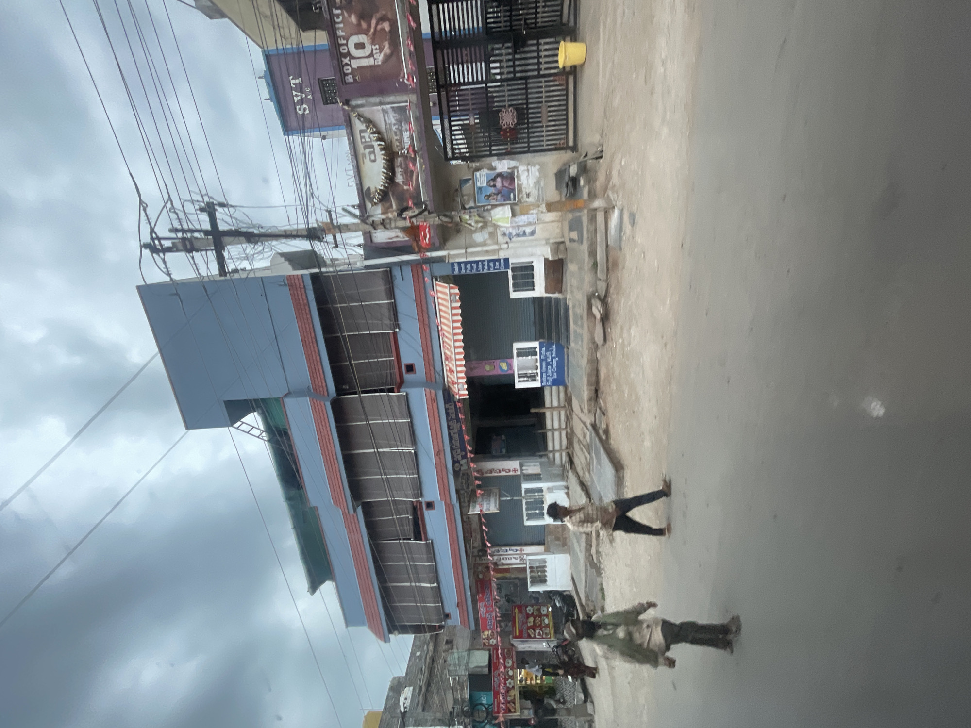 In Bethamcherla, India, the scene captures a street view with a multi-story building and its surroundings under a cloudy sky. The building, painted in blue and red, features multiple windows covered by blinds, suggesting commercial establishments on the lower floors. A sign with "SVT A/C" and "BOX OFFICE 10 DAYS" is visible on the right side of the building, indicating a cinema or entertainment venue. A metal gate is also present near this entrance.
In front of the building, two men are walking on the dusty street. One man, in the foreground on the left, wears a light-colored, possibly greenish, jacket and dark trousers. The other man, further to the right and slightly behind, is dressed in a checkered shirt and dark trousers. The street itself is unpaved and appears to be under construction or in a state of disrepair, with debris and wooden planks scattered about. A bright yellow bucket is placed near the gate.
The overall environment suggests a busy street in a developing urban area, with overhead power lines crisscrossing the sky, a common feature in many Indian cities. The weather appears overcast, contributing to a muted color palette of the scene.