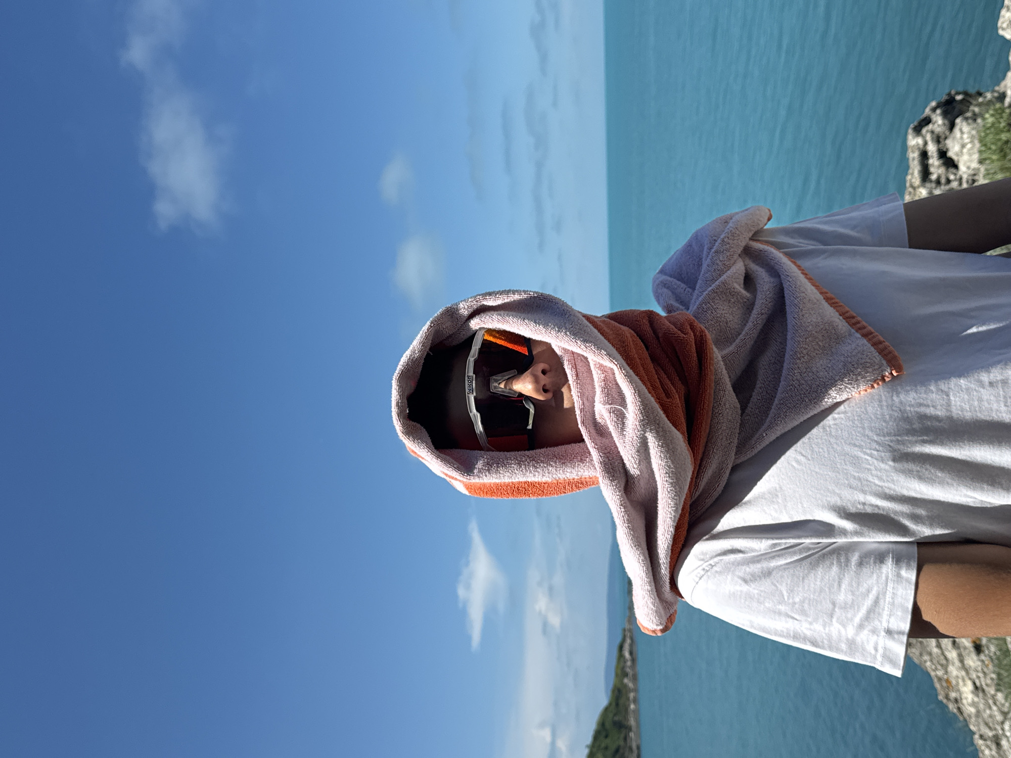 A person stands on a rocky outcrop overlooking a vibrant turquoise sea under a clear blue sky. The individual is wearing a white t-shirt and is partially covered by an orange and light pink towel draped over their head, obscuring their face except for their eyes, which are hidden behind large, white-framed sports sunglasses. The towel is wrapped around their head and shoulders, creating a hood-like effect. In the background, a distant green island or coastline is visible to the left, with the vast expanse of the sea stretching to the horizon. The sunlight casts shadows on the person's arm and clothing. The scene appears to be a sunny day at a coastal location, possibly in the vicinity of Tsarevo, Bulgaria, given the characteristic clear blue waters and rocky terrain often found along the Black Sea coast in that region.