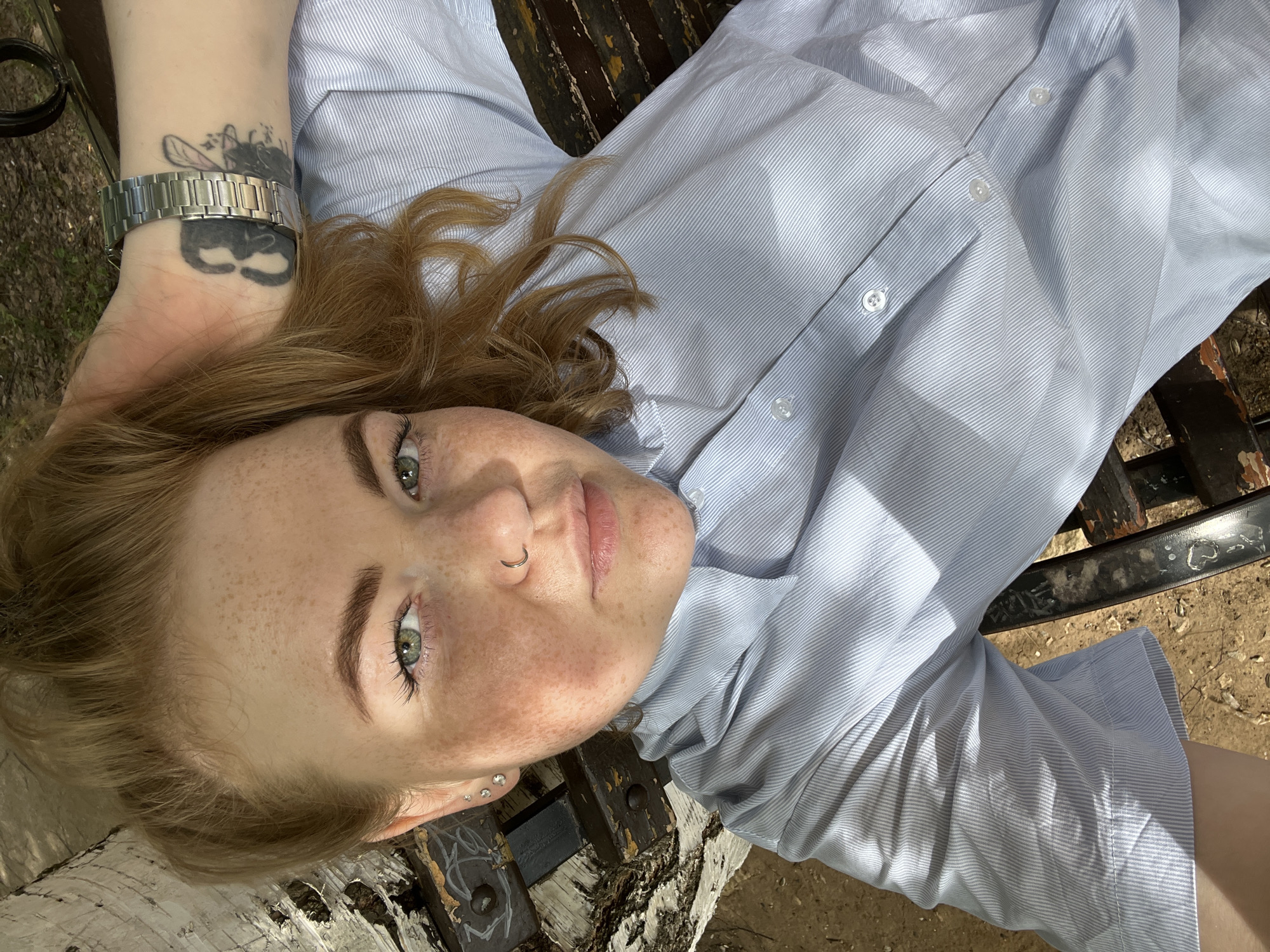 A young woman with freckles and reddish-blonde hair lies on a park bench in an outdoor setting. She is wearing a light blue striped button-down shirt and a metal watch on her left wrist. She has a tattoo on the same wrist. She has a nose ring and small earrings. The bench appears to be wooden with dark metal supports. The ground around the bench is earthy. The scene is lit by sunlight. The location is the city of Chaikovsky, Russia.
