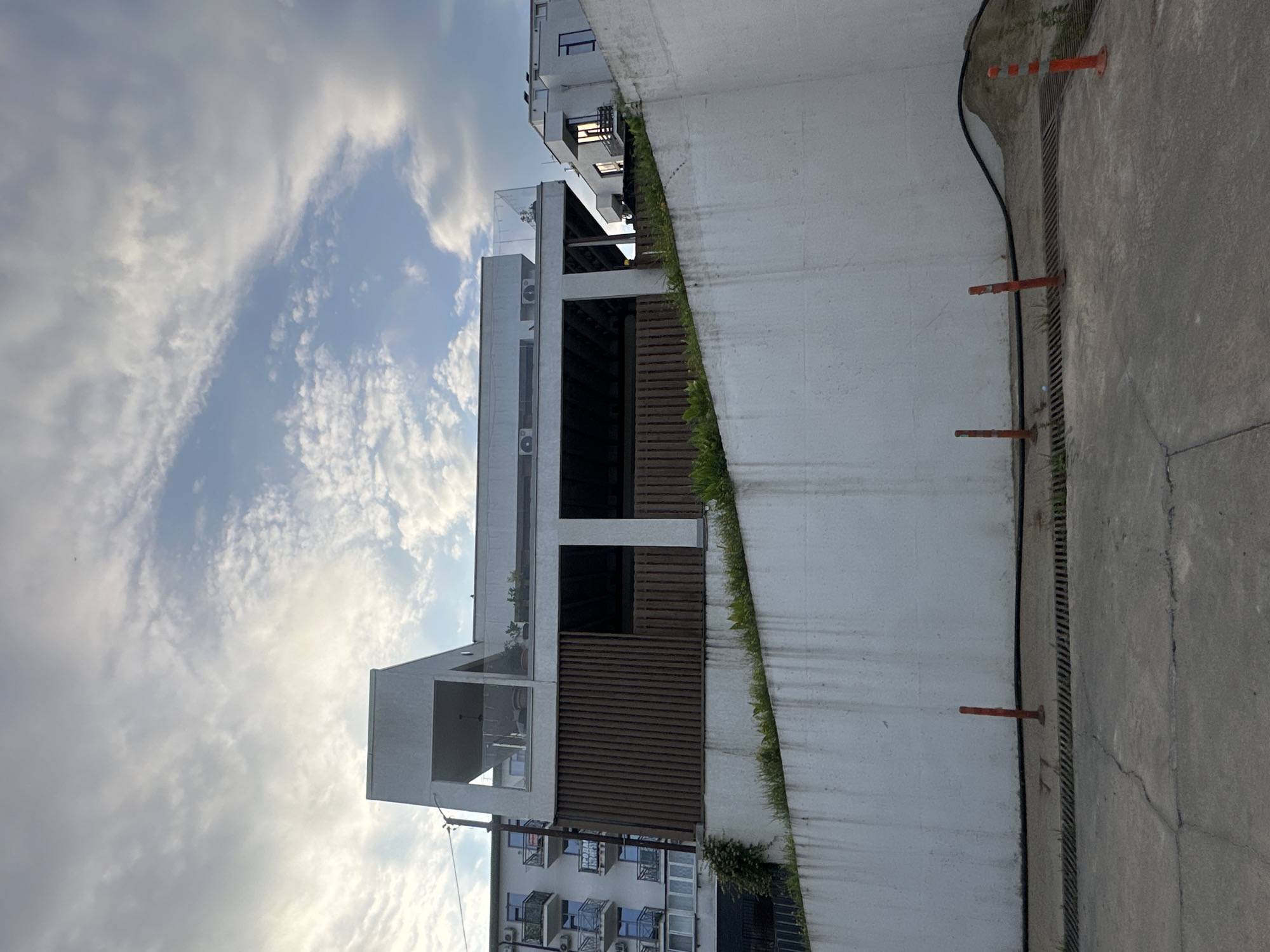 The image depicts a multi-story building with modern architecture, likely a hotel or apartment complex, situated on a hillside in Sochi, Russia. The building features a white facade with multiple balconies and large windows. A section of the building has a darker, slatted wooden exterior, and a prominent rooftop terrace with a glass railing is visible. The building is bordered by a steep, white retaining wall in the foreground, which is partially covered with grass and vegetation. In front of the retaining wall, a paved area with grates and several orange traffic cones is present. The sky above is partly cloudy, with patches of blue sky visible. The overall scene suggests an outdoor urban environment in a hilly coastal region.