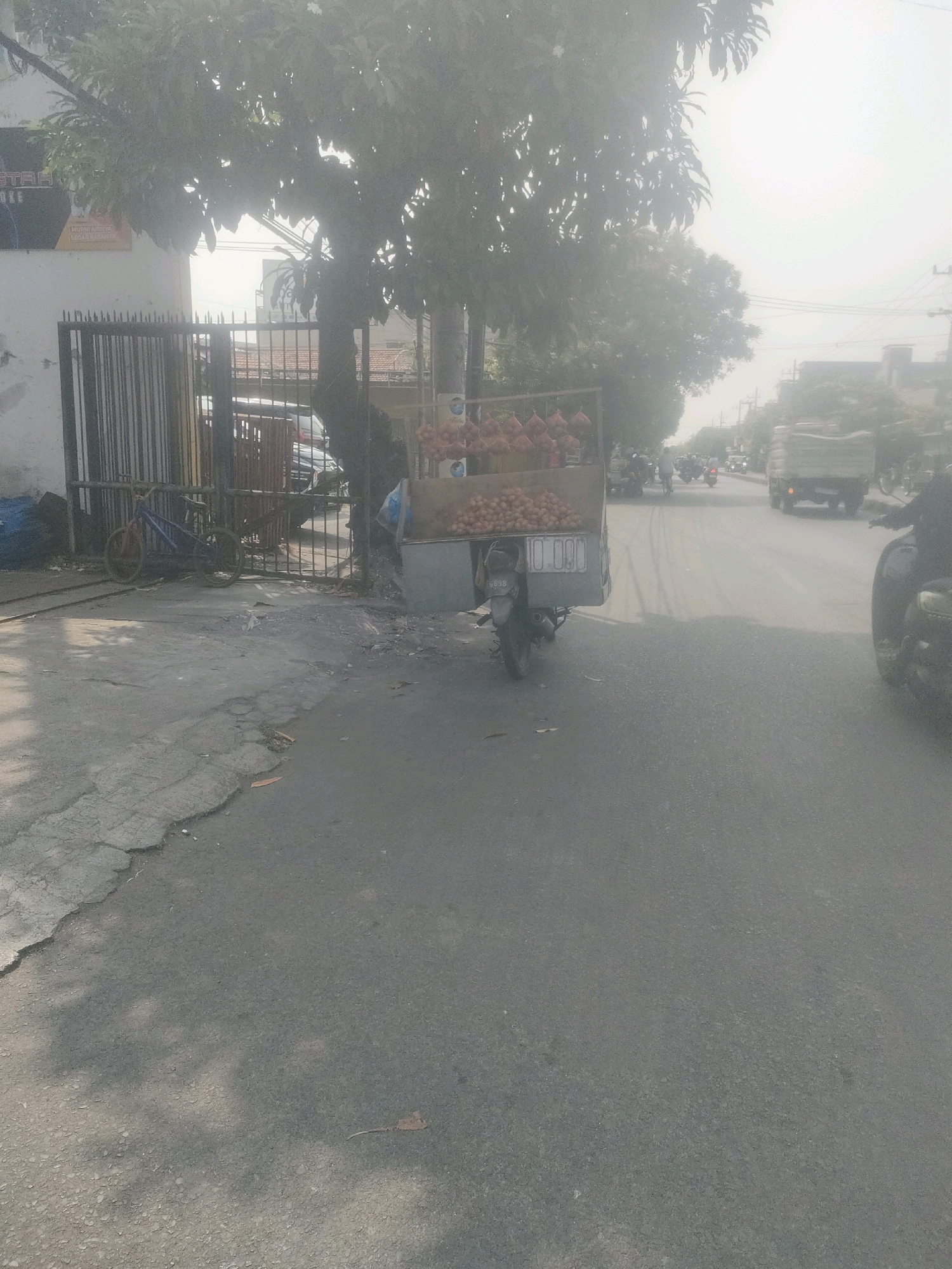 This is a street scene in Surabaya, Indonesia. The foreground shows a cracked asphalt road with shadows cast by trees. To the left, a metal gate stands in front of a building with signage that is partially visible. A blue bicycle is parked near the gate. In the middle ground, a small cart, likely a food or goods vendor, is parked on the side of the road. The cart is laden with what appears to be produce, possibly onions and potatoes. A motorcycle is attached to the back of the cart. Several other motorcycles and a truck are visible further down the street, indicating moderate traffic. The background is hazy, suggesting atmospheric conditions or dust.