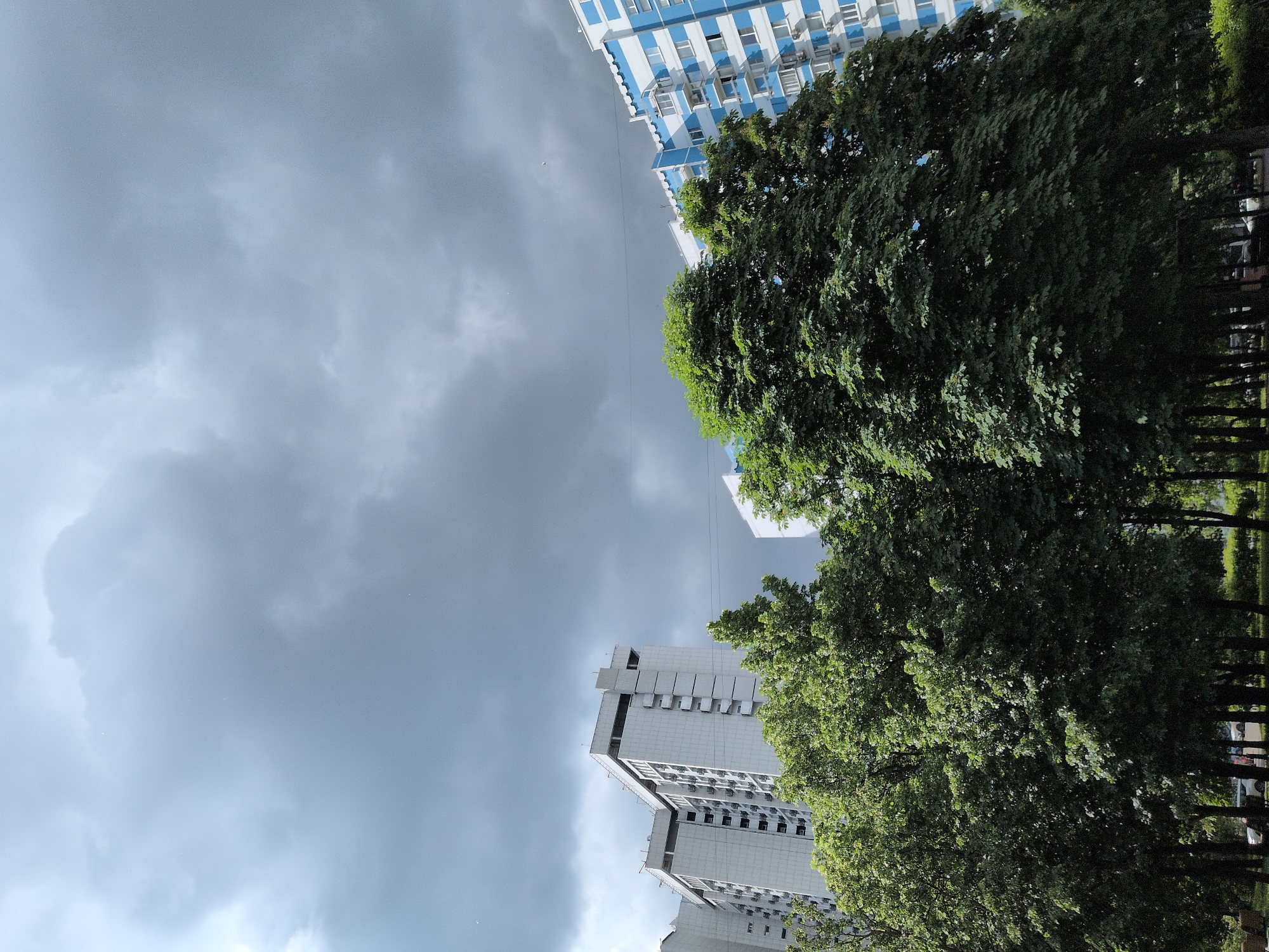 The image depicts an upward-facing view in Moscow, Russia. Tall buildings are visible on either side, one white and gray with visible window units and the other blue and white striped. Lush green trees are on the right and bottom edge. Above, a cloudy, overcast sky fills the center and left. No people are immediately visible in the image.