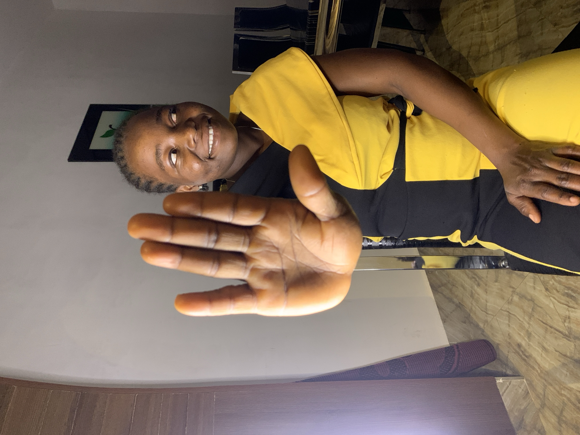 A woman is indoors, seated and facing the camera, her right hand extended towards the viewer. She is smiling and appears to be wearing a black and yellow garment. A framed picture hangs on the wall behind her. The location is likely a residence, given the flooring and furniture. The city of Jos, Nigeria, cannot be forensically confirmed from this image alone.
