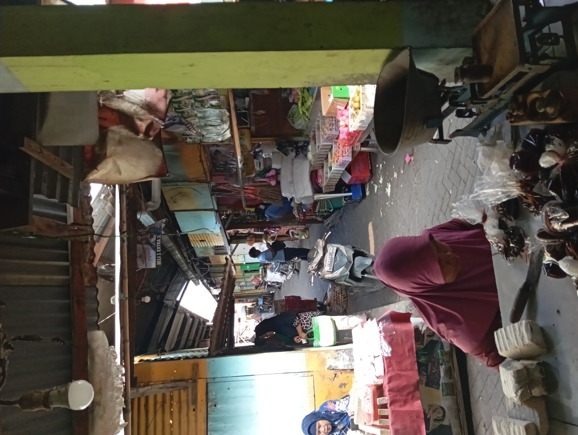 This image depicts a bustling market scene in Kepulungan, Indonesia. In the foreground, a person wearing a maroon hijab is seen interacting with items on a table, which include blocks of what appear to be tempeh, a knife, and several plastic bags filled with small dark objects, possibly spices or dried goods. To the left, a woman in a blue hijab is seated behind a table laden with goods, possibly food items.
The market stalls stretch into the background, covered by a corrugated metal roof. Various goods are displayed on shelves and tables, including produce like carrots and potatoes in crates. Several motorcycles are parked in the pathway of the market. People are seen walking and interacting within the market. The overall environment appears to be an outdoor, covered market, characteristic of many Indonesian cities. The lighting suggests it is daytime, with sunlight filtering through gaps in the roof. The presence of food stalls, diverse merchandise, and people engaged in commerce point to a typical day at a local Indonesian market.