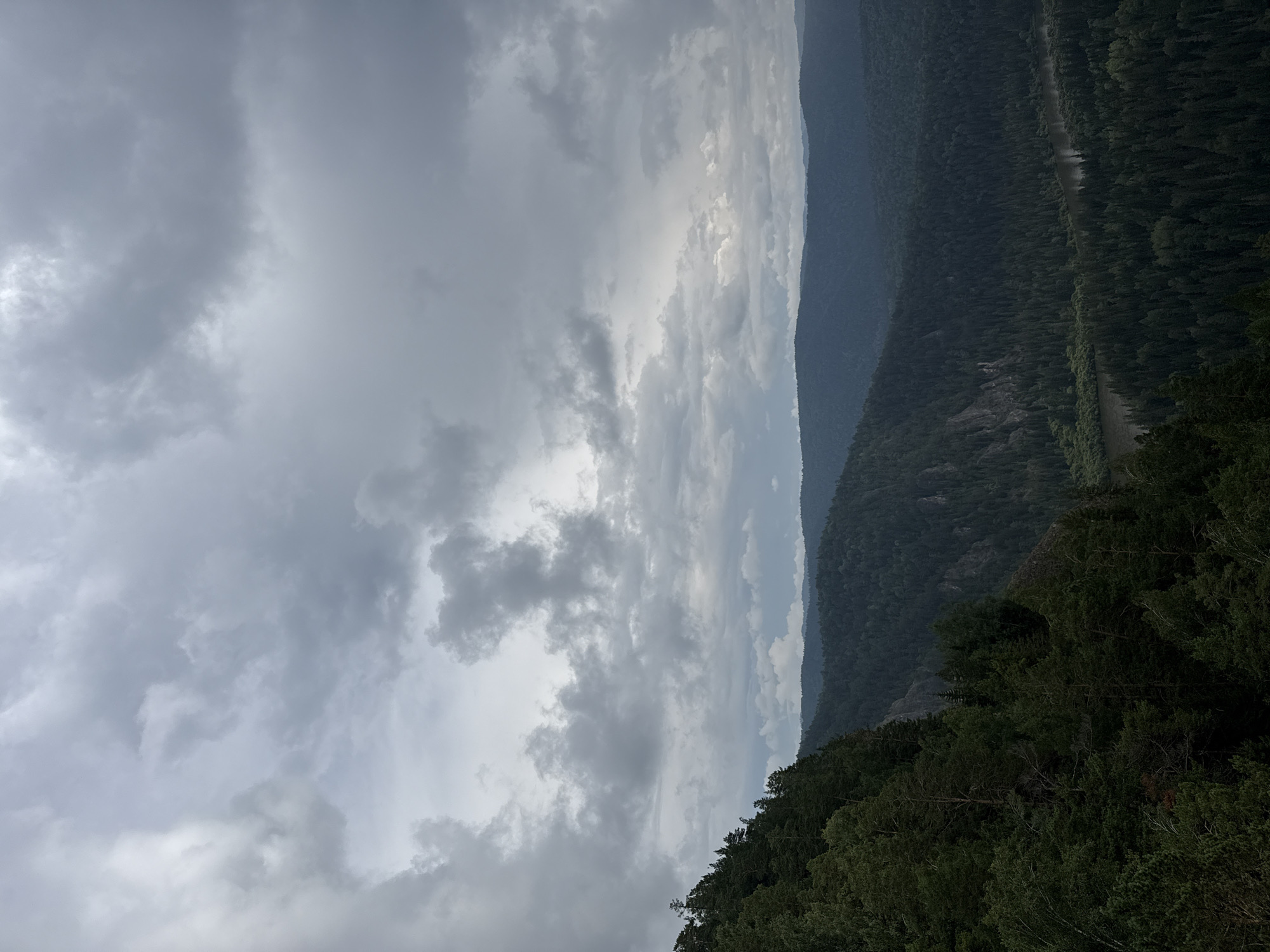 The image depicts a vast, forested mountain landscape under a heavily overcast sky. The foreground features a steep, densely wooded mountainside on the left, with dark green coniferous trees dominating the scene. In the middle ground, rolling hills covered in evergreen forests stretch into the distance, their contours softened by the atmospheric haze. A river meanders through the valley in the far distance, reflecting the muted light of the sky. The sky itself is filled with heavy, dark grey clouds, obscuring any direct sunlight and creating a somber, moody atmosphere. The image appears to be taken from a high vantage point, offering a panoramic view of the natural environment. The location is identified as Urman, Russia. There are no people, animals, or man-made structures visible in the image.