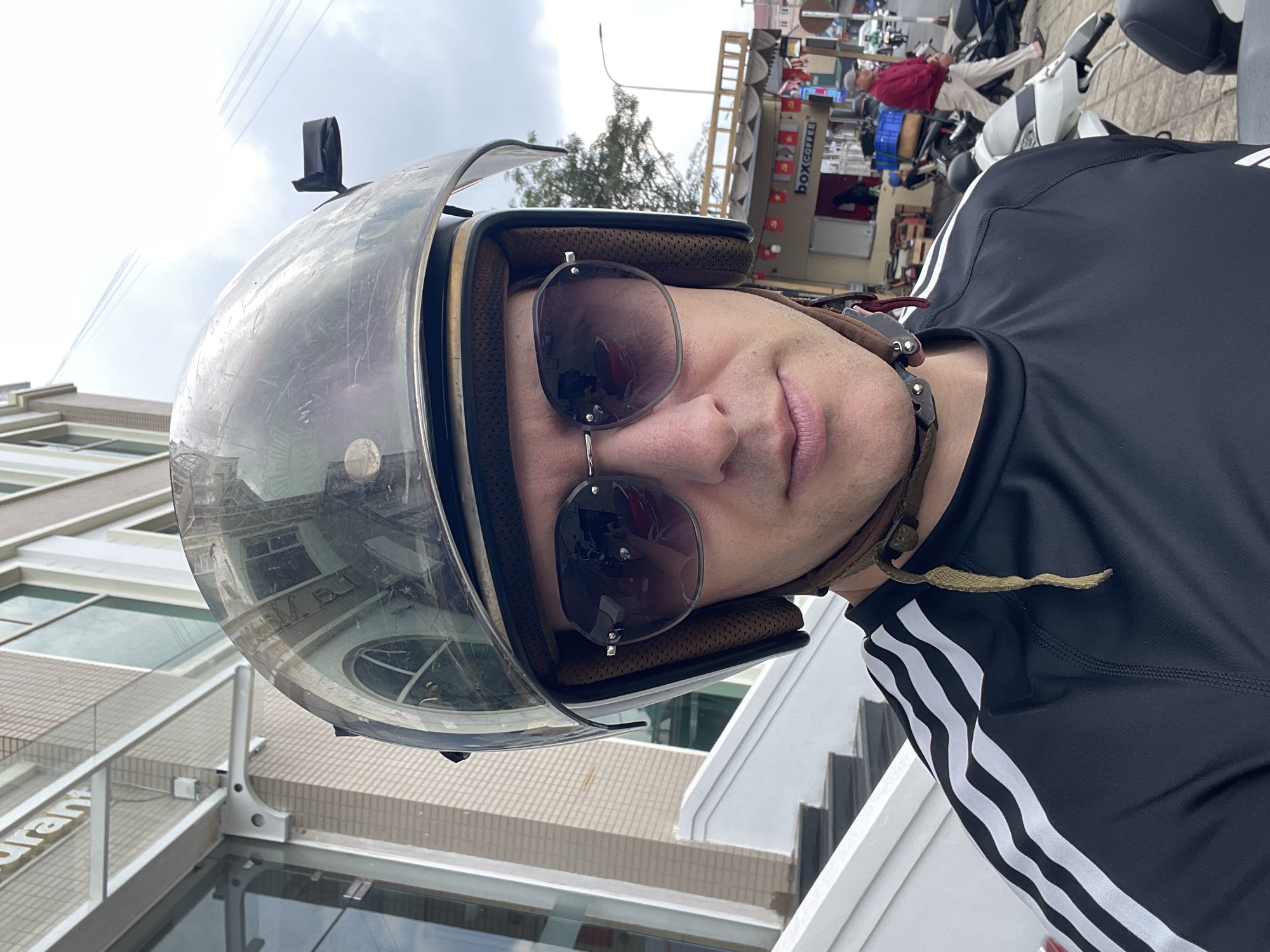 A medium shot captures a person wearing a motorcycle helmet and sunglasses. The helmet is silver with a clear face shield, and has a black mount affixed to the top. The sunglasses have dark lenses. The person wears a black long-sleeve shirt with white stripes on the sleeve.
The background indicates an urban environment. In the upper right, there is a parked moped and what appears to be a "BOX COFFEE" stand with Vietnamese flags. A building façade is visible in the lower-left. Reflections in the helmet's face shield reveal elements of the surrounding architecture. The location is inferred to be Lâm Viên - Đà Lạt, Vietnam.