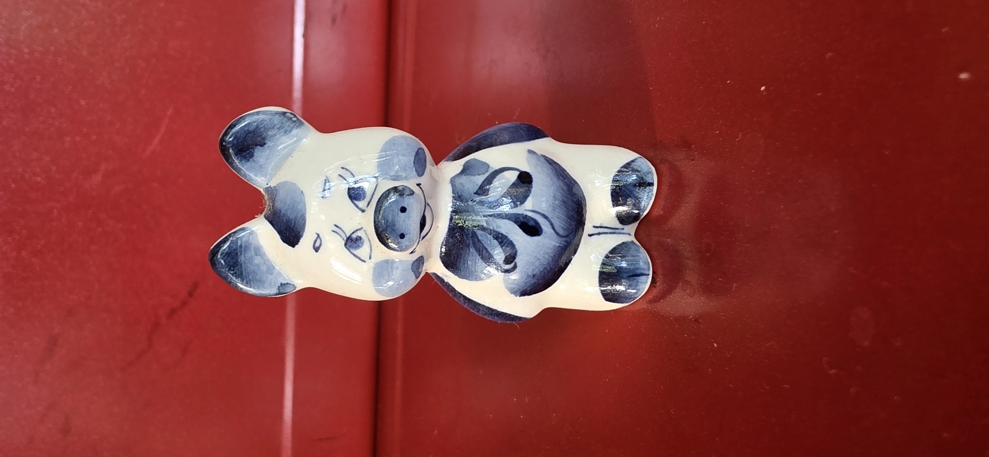 A small, white ceramic figurine of a pig with blue floral patterns is placed on a red, reflective surface. The pig has a rounded body, short legs, and large ears. Its face is adorned with blue painted eyes, eyebrows, cheeks, and a snout. A blue floral design is painted on its chest, resembling a bow. The background is a solid red surface with a slightly textured appearance. The lighting is even, casting a subtle reflection of the figurine onto the surface below. The scene is indoors, likely on a table or shelf. There are no people visible in the image.