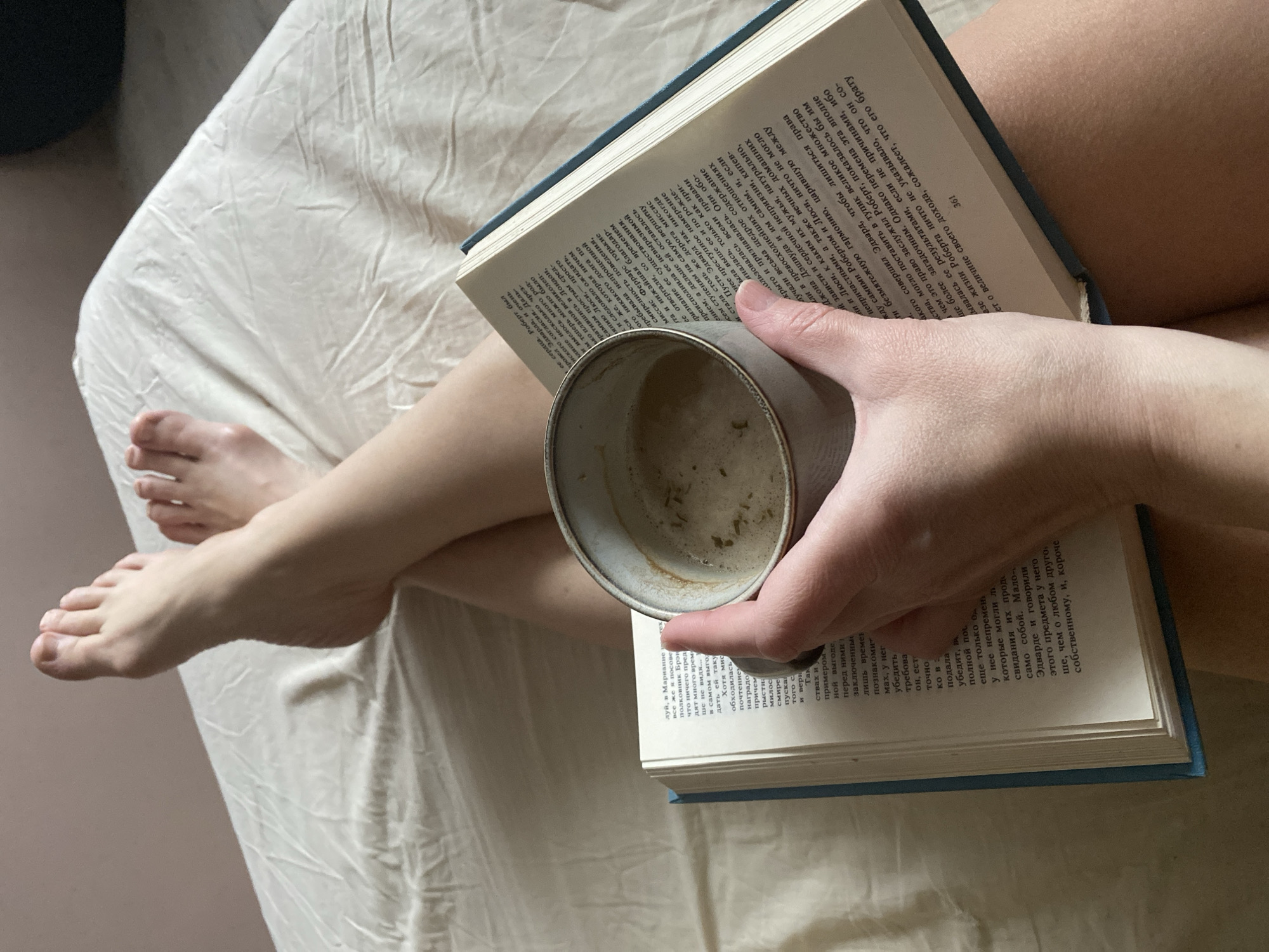 The image depicts a person lounging on a bed with a book and a cup of coffee. The person's legs are crossed, and their feet are visible on the left side of the frame. The book, held in the person's hand, is open, displaying text in a Cyrillic script (likely Russian), and has a blue cover. A grey cup, presumably containing coffee, rests on the open book. The bed is covered with a wrinkled, light-colored sheet. The overall setting suggests a relaxed, personal space, likely indoors. There is no way to establish the location based on this image.