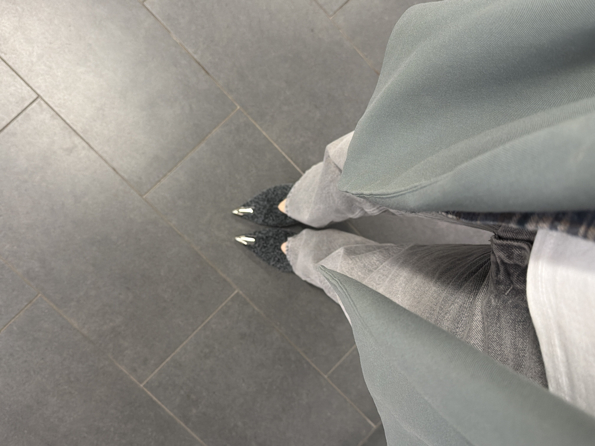 A first-person perspective photograph shows a person wearing gray jeans and shoes with pointed, metallic tips. The person is standing on a gray tiled floor. The lower half of a garment, possibly a coat or dress, of a matching muted sage green color, is draped to the right. The floor is composed of large, rectangular tiles arranged in a running bond pattern. The lighting is diffuse and uniform. The location is likely indoors due to the tiled flooring. No definitive markers within the photograph indicate this is in London, UK.