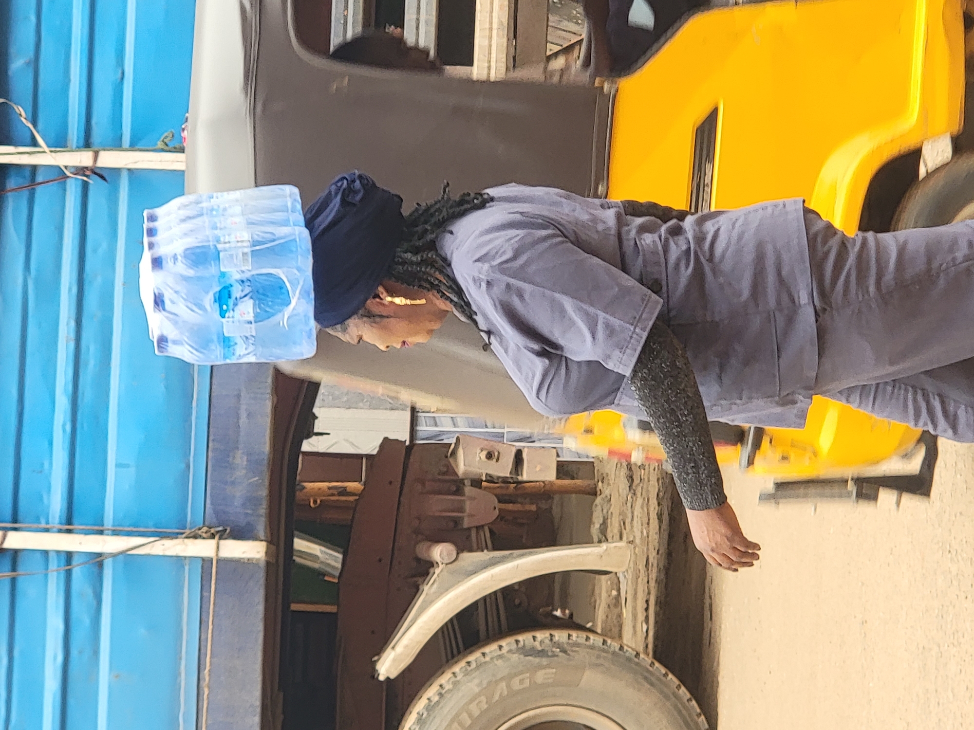 A person in a blue uniform, wearing a blue head covering and gold earrings, is carrying a stack of bottled water on their head in an outdoor location in the city of Maiburiji, Nigeria. The individual is standing next to a yellow truck and in front of a blue corrugated metal structure. The wheel of the truck is visible.