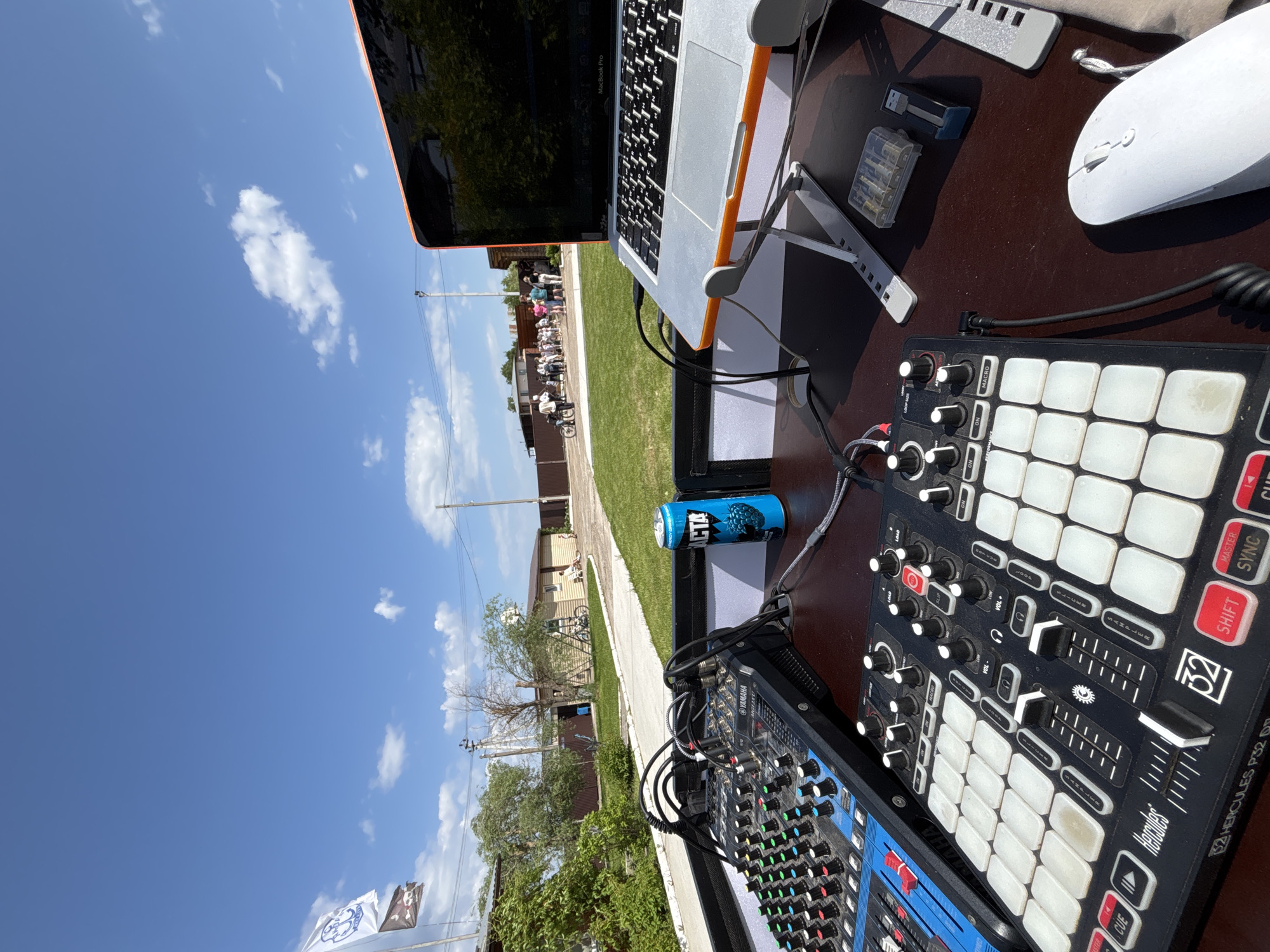 The image shows a DJ setup outdoors in Chaikovsky, Russia. A MacBook Pro laptop is elevated on a stand on a dark brown surface. Next to it is a white wireless mouse and a case of batteries. A blue can of "VICTA" energy drink sits near the laptop. To the right is a black Hercules P32 DJ controller. To the left is a blue Yamaha mixer. People are seen in the background walking along a paved path. The sky is blue with white clouds. Trees and grass are visible in the background. Banners or flags are seen on the lower left edge of the image.