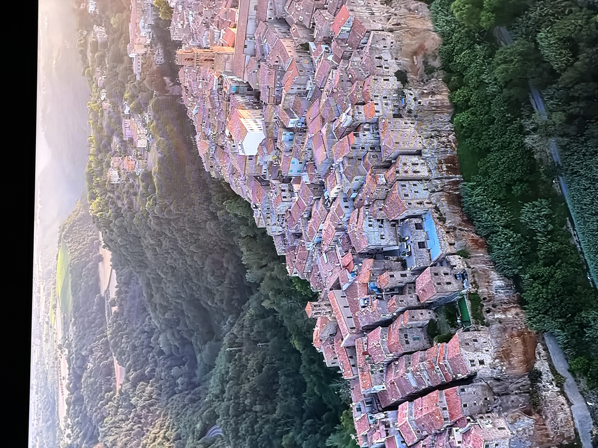 The image depicts an aerial view of a historic town built atop a sheer cliff face, overlooking a lush, green valley. The town is characterized by tightly packed buildings with terracotta roofs, many constructed from stone. A prominent bell tower rises from the center of the town. The valley below is densely forested, with a winding road visible in the distance. There are no visible people or specific actions depicted in the image, but the scene conveys a sense of ancient architecture and natural beauty. The question regarding the location in the city of Chirchik, Uzbekistan is not supported by the visual evidence in the image.