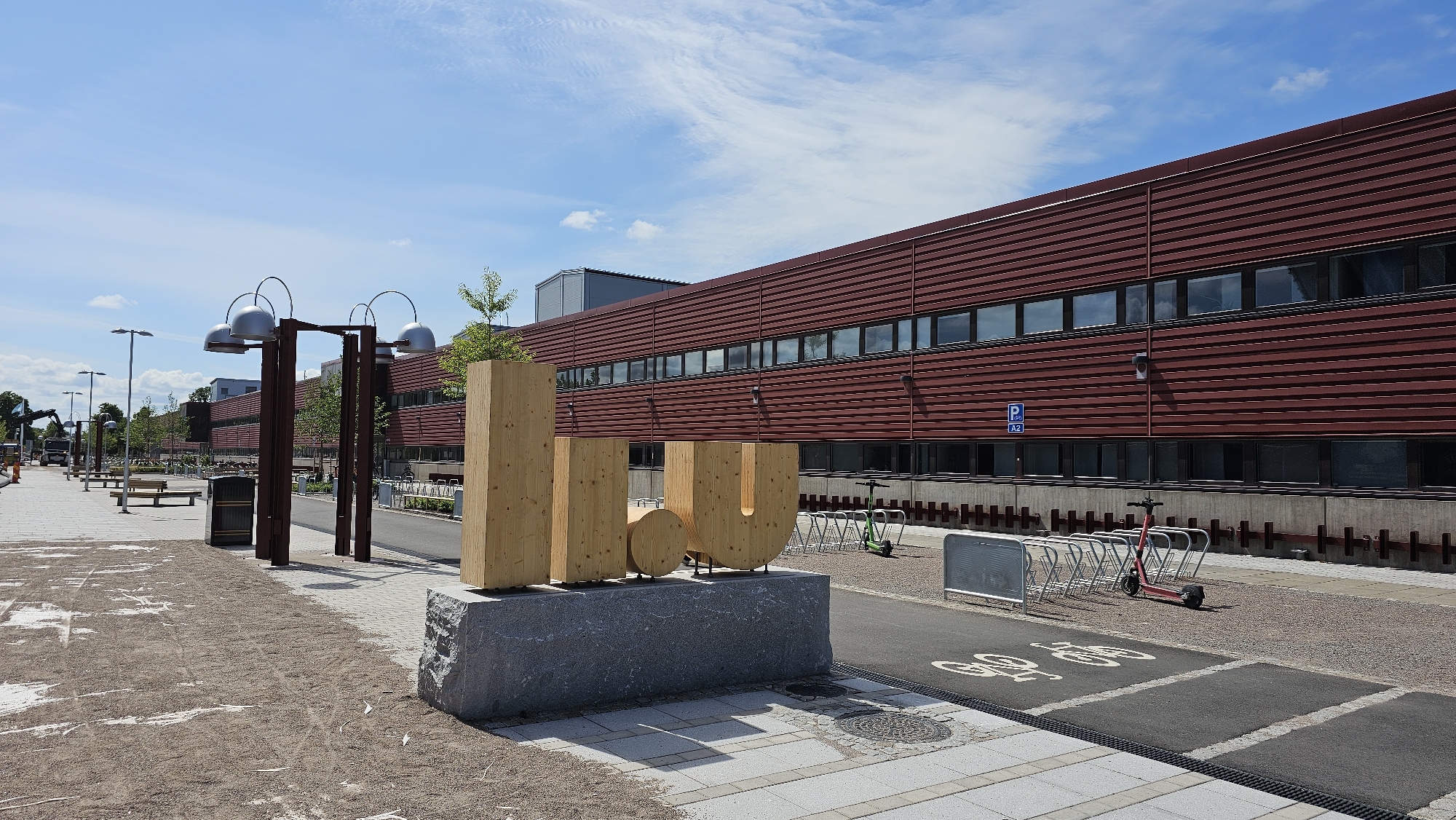 This is a wide, eye-level outdoor shot of the Linköping University campus in Linköping, Sweden, on a partly cloudy day. The main subject is a large, stylized wooden sculpture of the letters "LIU" (an abbreviation for Linköping University) resting on a granite base. Behind the sculpture is a long, modern building with a dark red, ribbed facade and a dark, continuous band of windows. To the left of the building and sculpture, a paved walkway stretches into the distance, lined with benches, trash receptacles, and street lamps. The walkway transitions to a gravel area in the foreground. A road is visible to the right of the sculpture, featuring a bike lane marked with cycle symbols, a row of empty bike racks, and a red electric scooter parked by the racks. In the background, more campus buildings and trees are visible under a bright blue sky with wispy white clouds. A construction vehicle is partially visible on the left side of the walkway in the distance.
