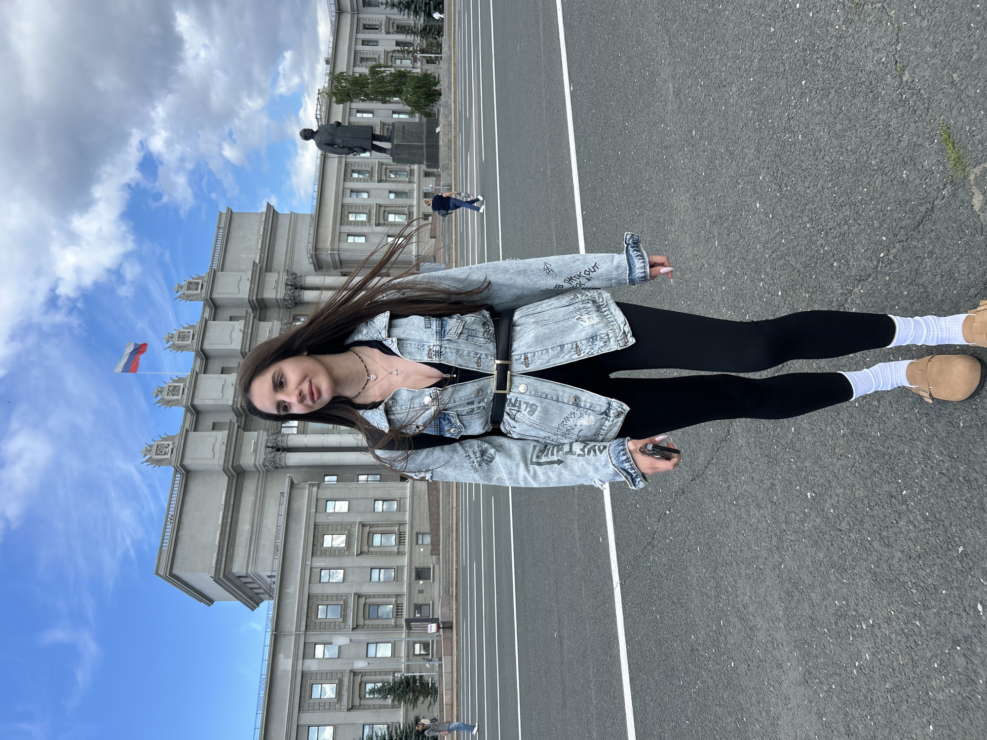 A young woman with long dark hair is standing in front of a large, imposing building. She is wearing a denim jacket, black leggings, white socks, and light brown shoes. The jacket has graphics on the sleeves. She is holding a cell phone in her right hand. She is looking towards the camera with a slight smile.
The building behind her appears to be made of stone or concrete and has many windows. A Russian flag flies from a pole in front of the building. To the left of the woman, a statue of a man in a long coat and hat stands on a pedestal. In the background, other people can be seen walking in the distance.
The scene is an outdoor public space, likely a plaza or square, with an asphalt surface marked with white lines, suggesting it may be a road or walkway. The sky is blue with scattered white clouds, indicating daytime weather. The overall atmosphere is neutral, with the woman appearing relaxed and confident. The location is identified as Novokuybyshevsk, Russia.