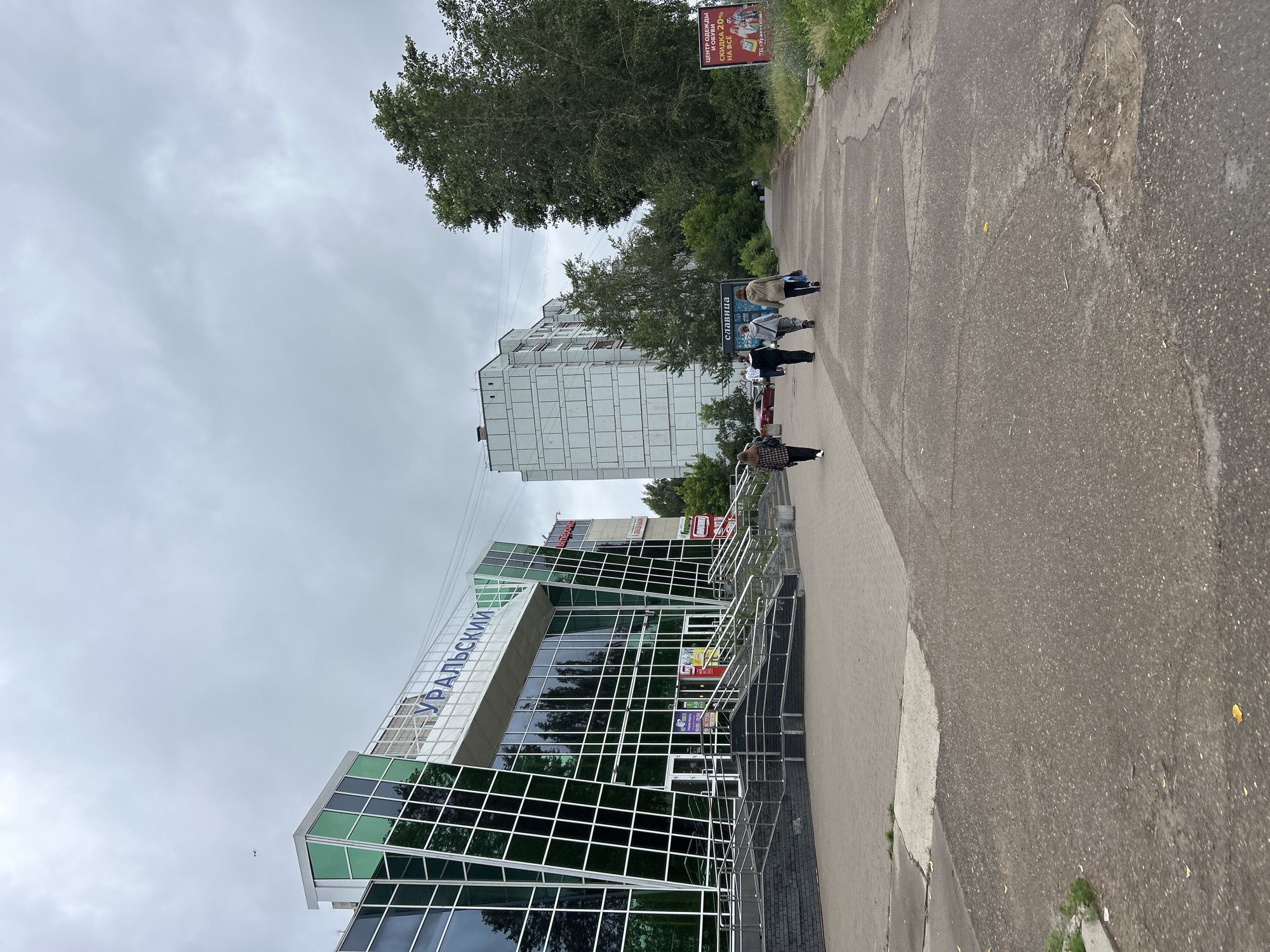 The image depicts a scene in the city of Chaikovsky, Russia, focusing on a modern building with a distinctive green glass facade and the signage "УРАЛЬСКИЙ" (Uralskiy). In front of this building, a paved walkway leads towards a large, light-colored apartment building in the background. Several people are walking away from the viewer on this walkway. To the right of the walkway, a tall, mature tree stands prominently, and a billboard advertising clothing and shoe sales with a "СКИДКА 20% НА ВСЁ" (20% discount on everything) message is visible. The sky is overcast and gray, suggesting a cloudy day. The overall environment appears to be an urban or suburban setting.