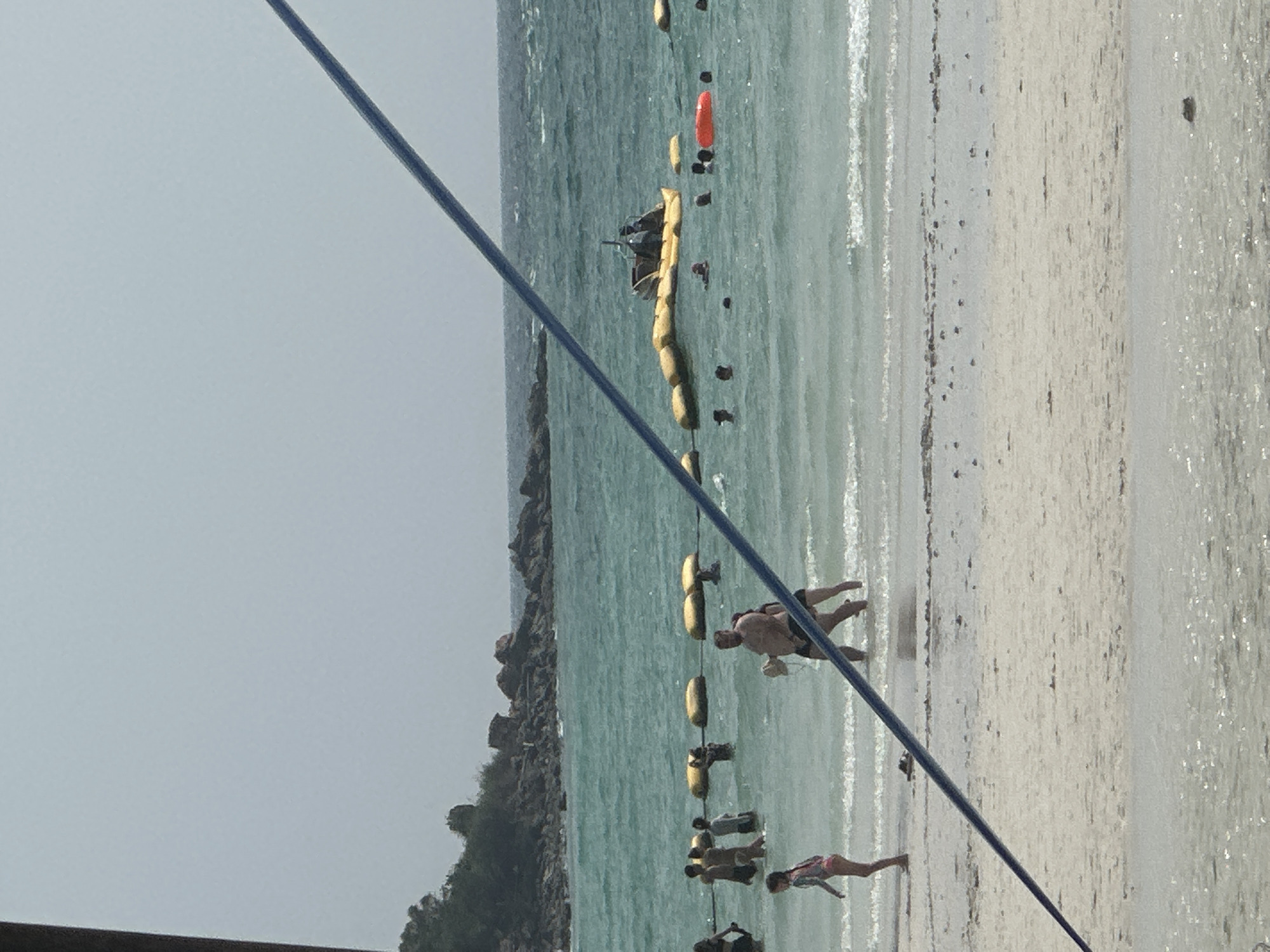 The image depicts a beach scene, likely in Pattaya City, Thailand, given the location provided. The view is from an elevated position, possibly a building or structure near the beach, partially obscured by a blue cable or rope in the foreground.
In the midground, the turquoise sea meets a light-colored sandy beach. A series of yellow buoys and what appear to be tethered rafts or platforms are visible in the water, with a few people swimming or standing nearby.
On the beach, several individuals are scattered along the shoreline. Some are swimming, while others are walking along the sand. The background includes a rocky coastline with some vegetation. The sky is overcast, creating diffused lighting.