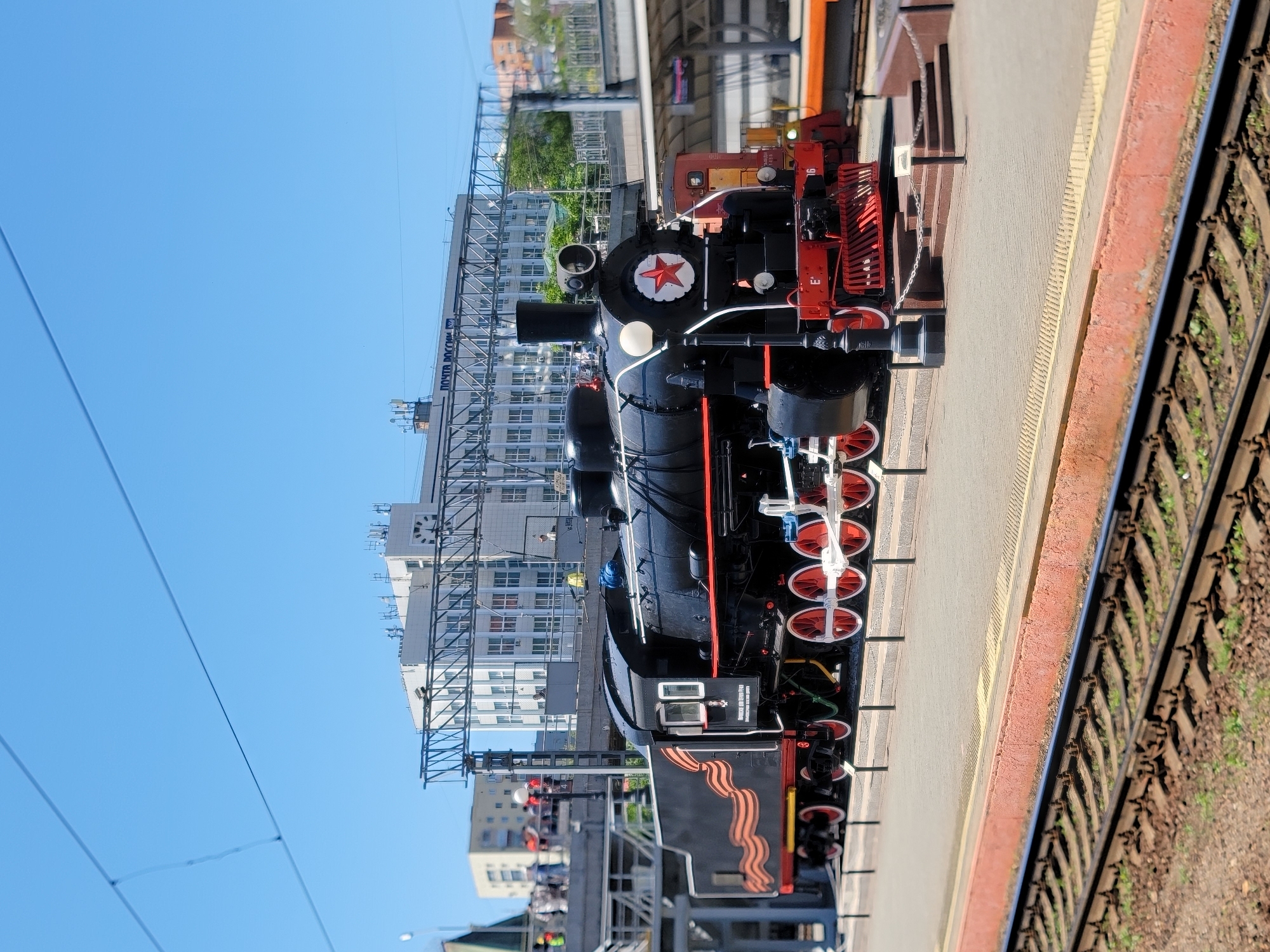 Here's a detailed description of the image:
**Scene Overview:**
The image depicts a black steam locomotive, identifiable by a red star emblem, positioned on a train platform in an urban setting. Railway tracks run adjacent to the platform.
**Objects and Details:**
* **Locomotive:** The engine is primarily black with red detailing, including the wheels, some of the framework, and decorative elements. The red star emblem is prominently displayed on the front of the engine. The locomotive features multiple wheels, connecting rods, and other mechanical components typical of steam engines.
* **Train Platform:** The platform is made of concrete or paving stones, and it is bordered by a red painted edge.
* **Railway Tracks:** Parallel railway tracks are located next to the platform, with the rails showing signs of rust and wear.
* **Buildings and Infrastructure:** High-rise buildings are visible in the background, suggesting an urban or suburban location. Overhead power lines and supporting structures are also present.
**Environment:**
* **Weather:** The sky is clear and blue, suggesting sunny weather conditions.
* **Location:** The presence of railway tracks, the platform, and the surrounding buildings indicates a train station or railway museum setting in a city.
**People:**
* There are no people visible in the immediate foreground. However, some blurred figures can be seen on the platform behind the train.