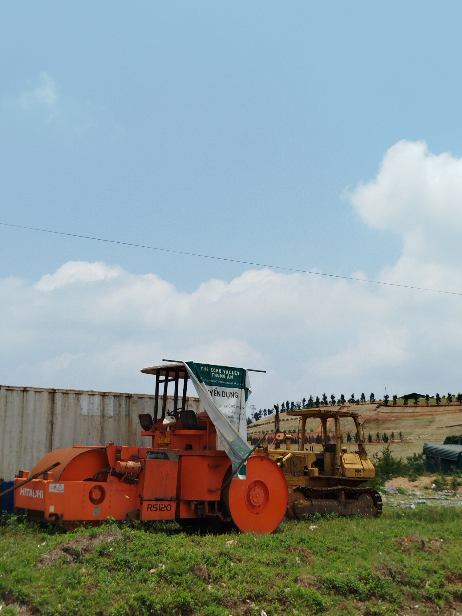 In Đa Nghịt, Vietnam, a partially obstructed view shows a construction site with various heavy machinery. An orange Hitachi RS120 road roller sits beside a yellow D41A bulldozer, partially obscured by a green and white advertising banner reading "THE ECHO VALLEY THUNG ÂM" (The Echo Valley Thung Am). The banner lists job openings: "YEN DUNG" (Seeking), and the categories "CHUNG MAN," "NHANVENC VU," and "PVE."
A shipping container acts as a backdrop to the vehicles. Behind them, a terraced hill has evenly spaced trees planted, suggesting landscaping. The sky is mostly clear, with scattered clouds and a high-voltage power line cutting across the scene. The machinery sits on a grassy area.