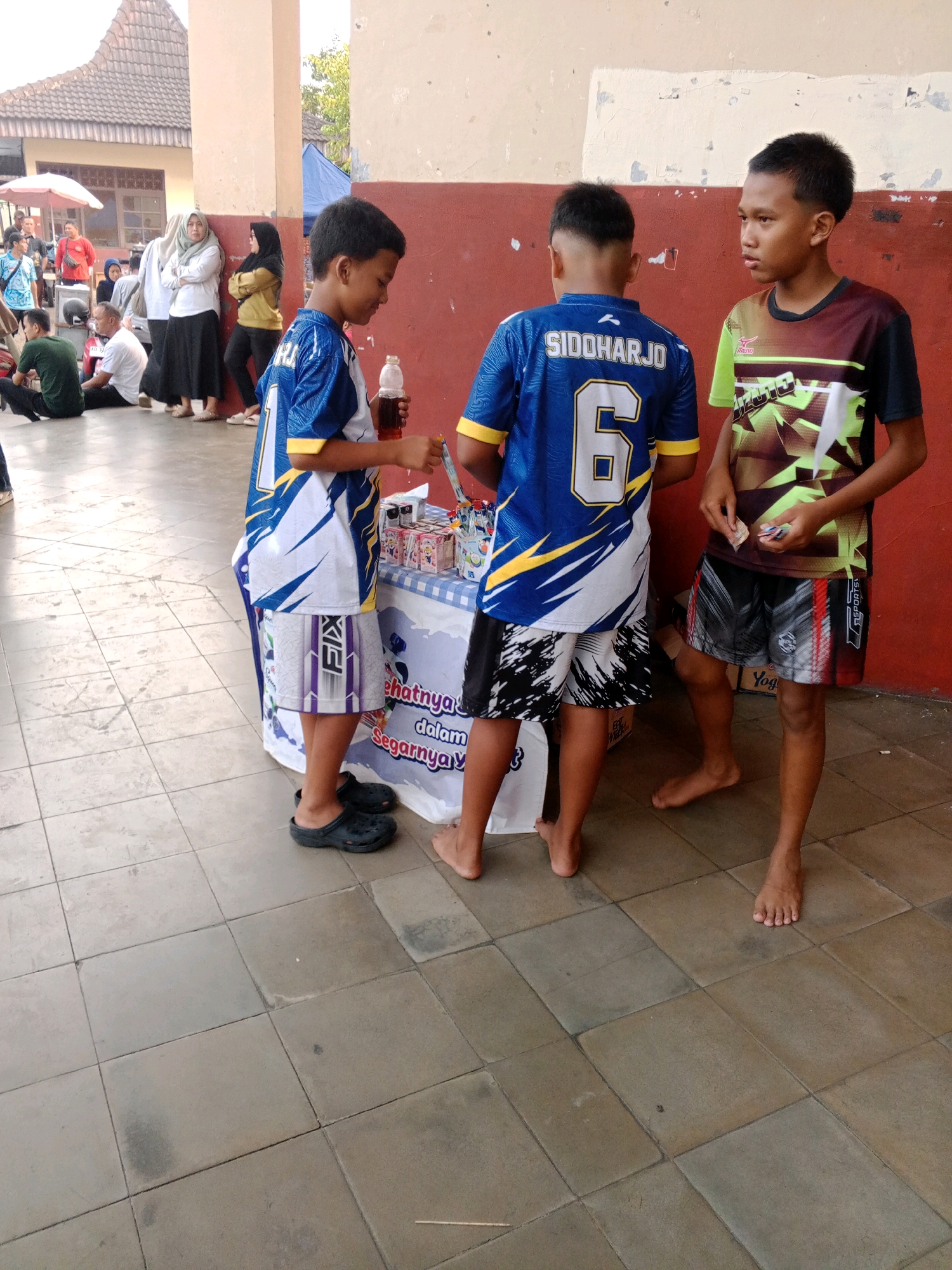 Three boys are standing around a table with a blue and white checkered cloth in Wuryorejo, Indonesia. The boy on the left, wearing a blue jersey with "SIDOHARJO" and the number "6" on it, is holding a clear bottle of dark liquid and a small object. He is wearing black sandals. The boy in the middle, also wearing a similar blue jersey and patterned shorts, is looking down. The boy on the right, wearing a colorful patterned jersey and black shorts, is holding a few small objects in his hand. The table is laden with various small items, including boxes of what appear to be drinks. In the background, there are other people, buildings, and trees, suggesting an outdoor public space. The ground is tiled with square grey tiles.