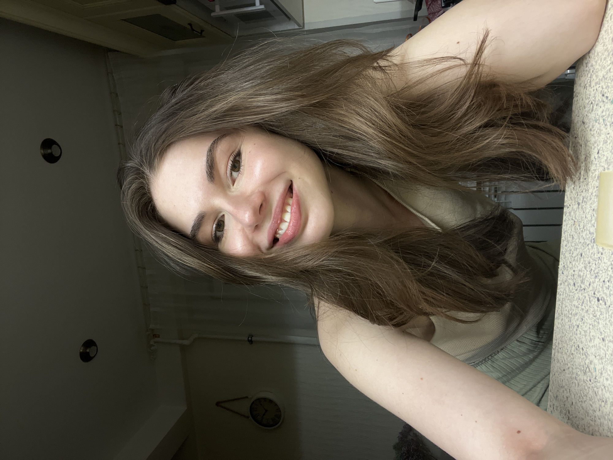 A young woman with long brown hair smiles into the camera. She has light skin, brown eyes, and thick eyebrows. Her teeth are visible, and she appears to be wearing lip gloss. She is wearing a beige top. The background appears to be an interior, possibly a kitchen or bathroom, given the presence of a countertop, wall-mounted clock, and light switches. The location in Saint Petersburg, Russia, is not discernible from the image.