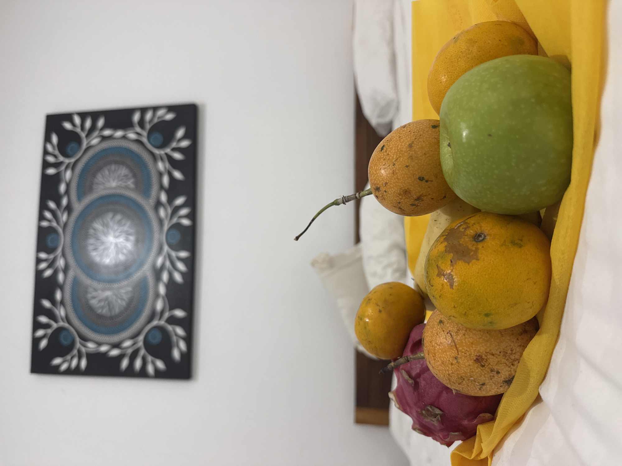 A still life composition is captured indoors with a focus on a collection of fruit and an artwork. On the left, an art piece hangs on a white wall, featuring a black background with concentric circular designs in shades of blue and gray, surrounded by a decorative white border. To the right of the artwork, a bundle of fruits is arranged, including green apples, passion fruit, oranges, a mango, and a dragon fruit. These fruits are held within a white and yellow cloth, adding layers of color and texture to the composition. The location, Kuta, Indonesia, is not evident from visual cues in the image.