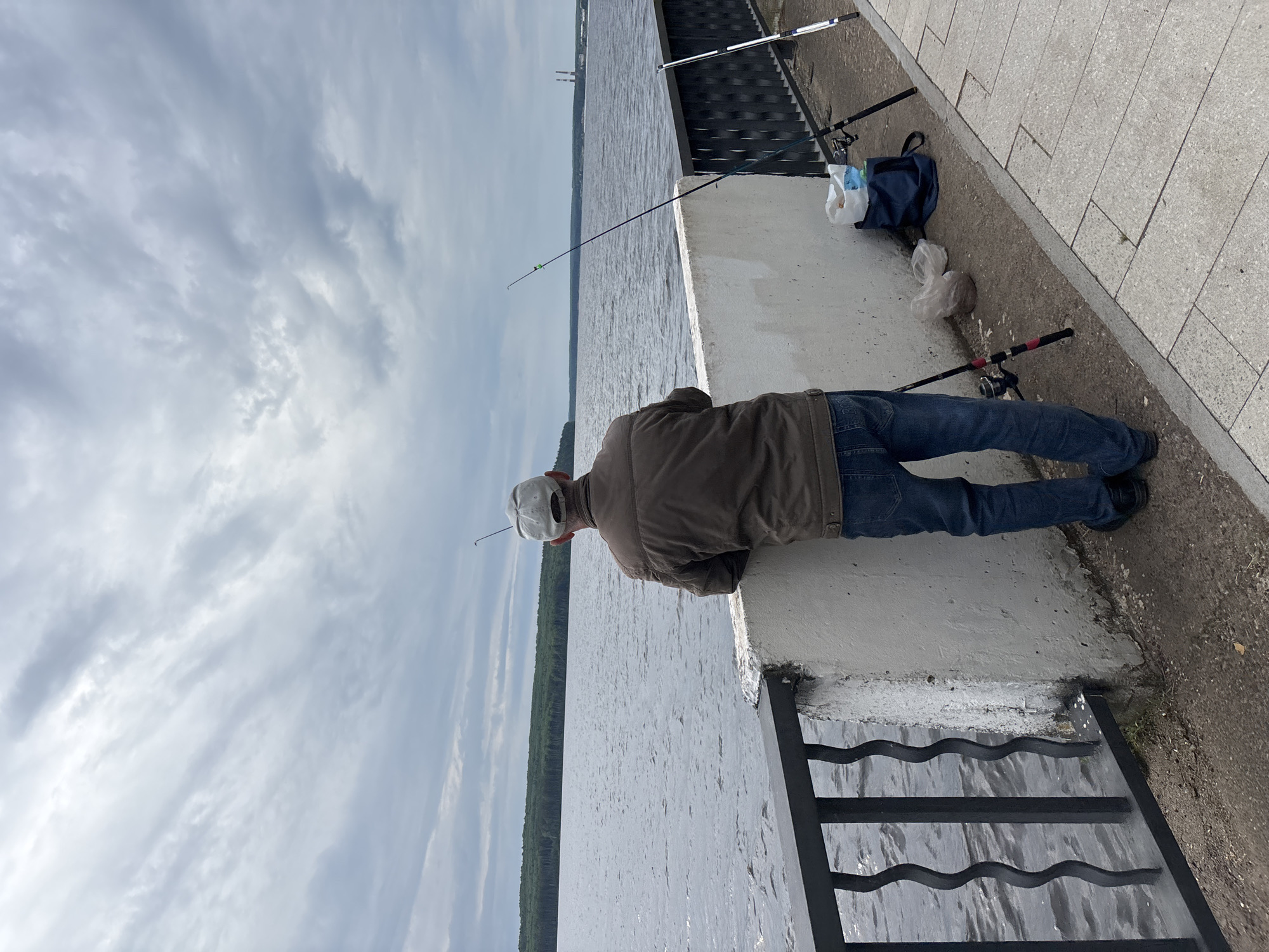 This media file captures a man fishing from a concrete embankment on an overcast day in Chaikovsky, Russia. The image is taken from behind the man, showing him leaning over a low concrete wall, looking down at the water. He is wearing a brown jacket, blue jeans, and a white baseball cap.
Several fishing rods are visible on the embankment. One rod extends over the water near the man, while at least two others are set up further along the concrete edge, resting on the ground. A dark blue bag, a white plastic bag, and a brown plastic bag, likely containing fishing supplies or personal items, are placed on the paved walkway next to the fishing rods. The ground comprises concrete paving stones and a dirt/gravel strip along the edge.
The scene features a wide body of water, possibly a river or large lake, stretching far into the distance. A green treeline marks the opposite bank, and in the very far distance, some industrial structures, resembling chimneys or cooling towers, can be discerned. The sky is largely covered with light grey, streaky clouds, indicating an overcast but bright daytime. A black metal railing or fence is visible in the foreground, partially obscuring the lower right portion of the image. There is no visible text in the image.