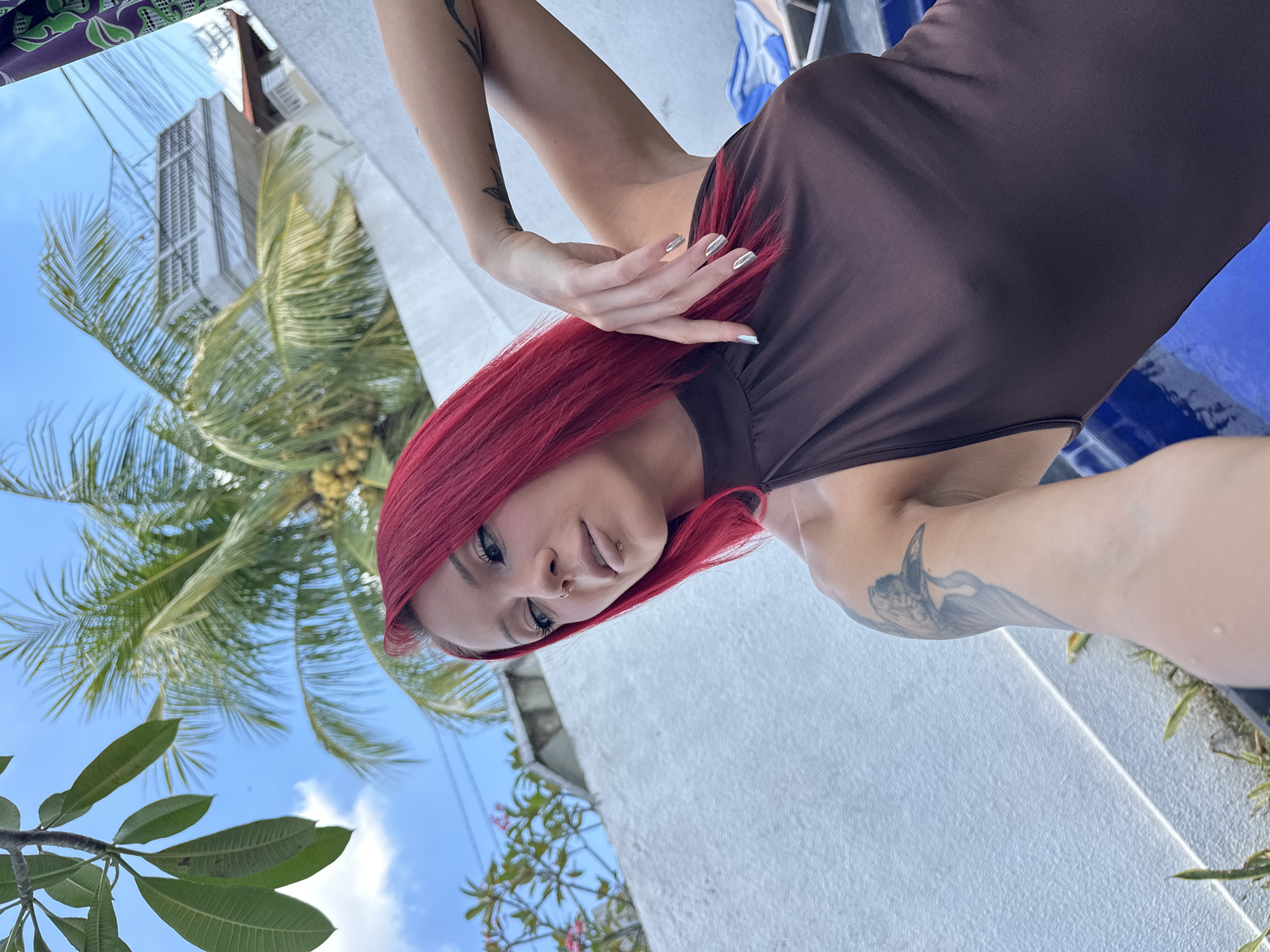 A woman with red hair, fair skin, tattoos on her left arm, and facial piercings (nose and lip) stands in an outdoor environment with tropical vegetation, likely in North Kuta, Indonesia. She is wearing a brown sleeveless dress and has silver nail polish. The background includes a building, palm trees, and a blue sky with some clouds. She is holding her hand near her face and looking at the camera.