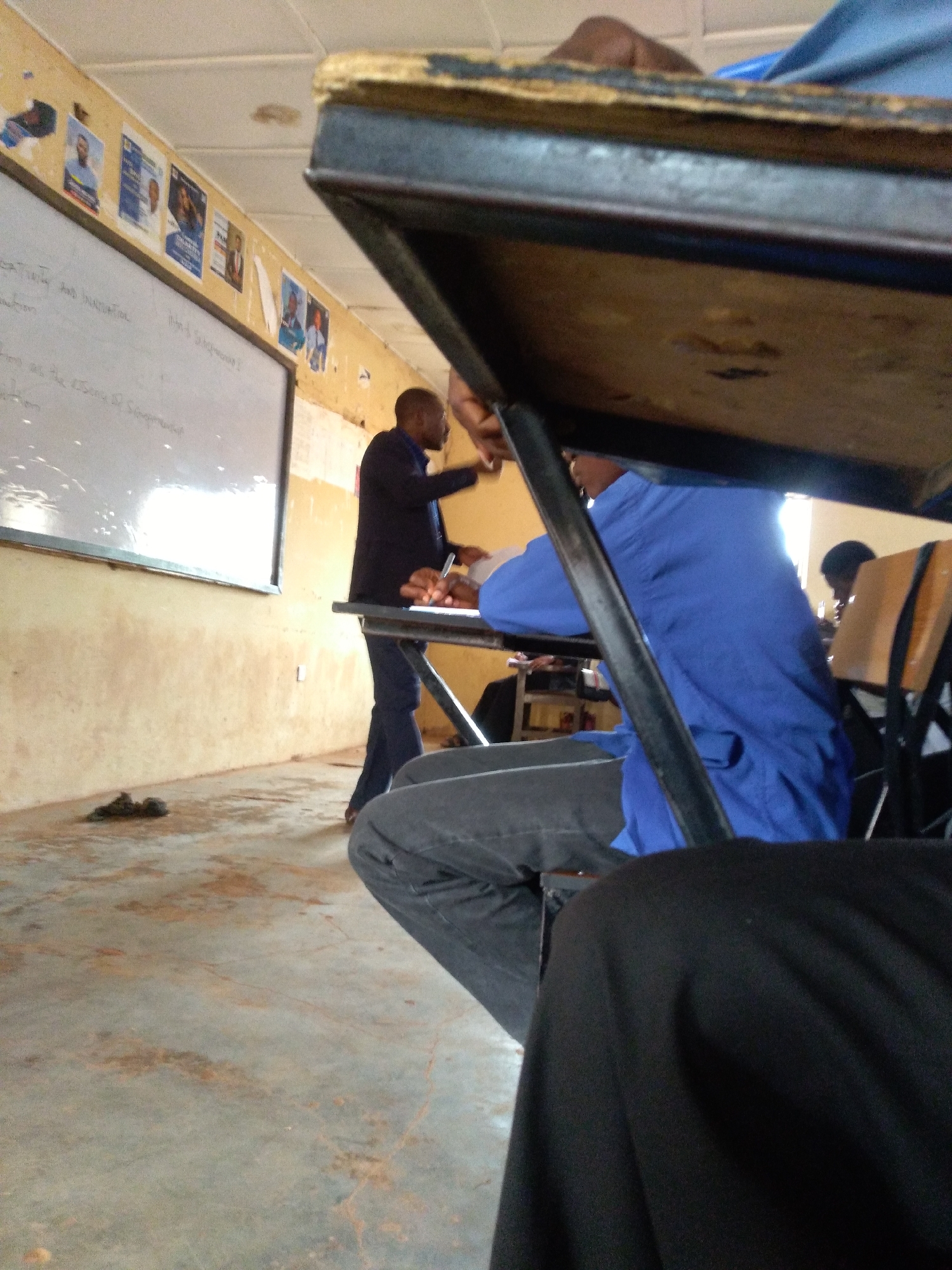 The image depicts a classroom scene in Jos, Nigeria. A man, presumably a teacher, stands near a whiteboard, gesturing with his hands. He is wearing a dark suit jacket over a blue shirt and dark trousers. The whiteboard has some text written on it, partially obscured. Along the wall above the whiteboard, several posters are displayed, some featuring portraits of individuals. The classroom appears sparsely furnished, with a few desks visible. In the foreground, parts of a student are visible, seated at a desk and wearing a blue garment. The floor is concrete and shows signs of wear and some stains. The overall impression is of an active learning environment in a modest educational setting.
