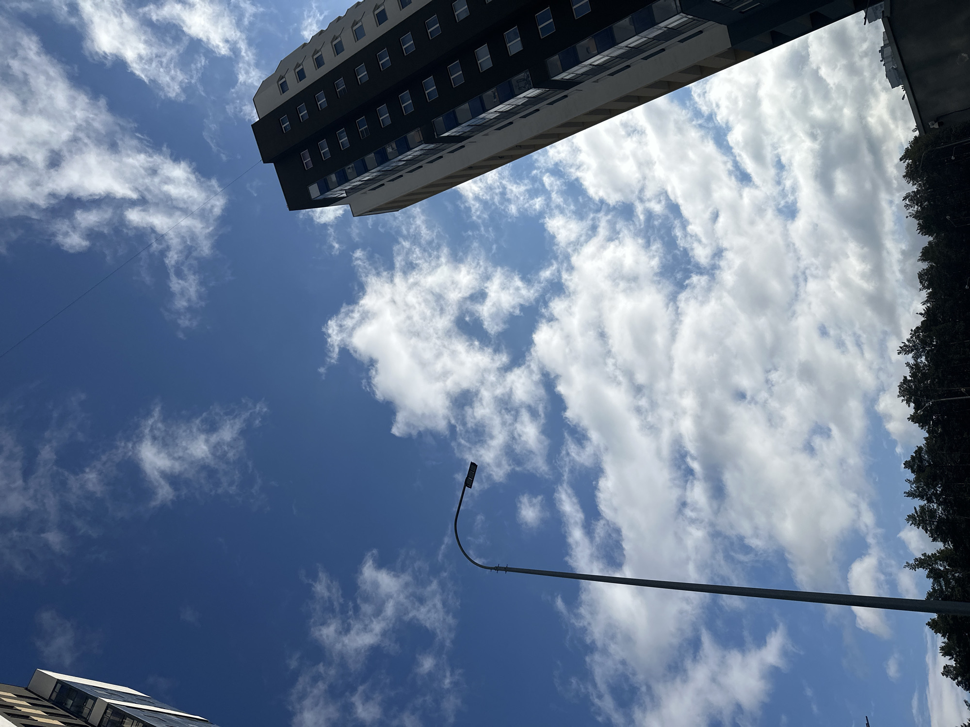 The image shows a low-angle view of the sky with scattered clouds and buildings in Yekaterinburg, Russia. A tall building with multiple windows is visible in the upper left corner. A street light post is also in the frame. Trees are seen at the right edge of the image. Another building is partially visible at the bottom left corner.
