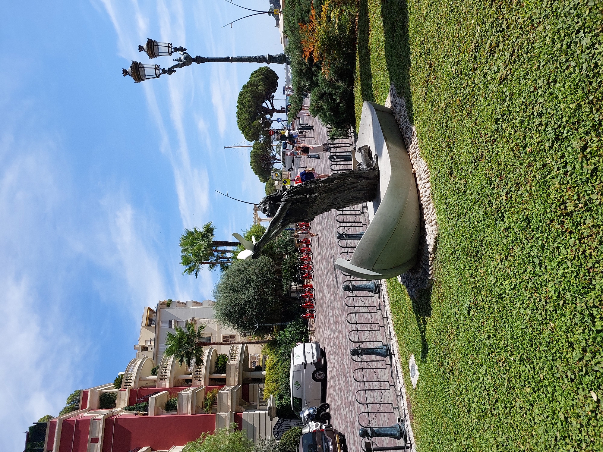 The image depicts a sunny day in Monaco, with a clear blue sky and scattered white clouds. In the foreground, a sculpture of a person in a boat is situated on a green lawn. The sculpture appears to be made of bronze and is weathered, with a figure reaching upwards. The boat is a light gray color, and it rests on a bed of white pebbles.
Behind the sculpture, a black metal fence separates the lawn from a paved walkway. On the walkway, several people are visible, some walking and others standing. To the left of the walkway, a line of red bicycles is parked, suggesting a public bike-sharing service.
Further in the background, a white van is parked, and several other vehicles are visible on the street. Buildings with red and white facades, characteristic of Monegasque architecture, rise above the street. Palm trees and other greenery dot the landscape, adding to the Mediterranean feel of the scene. A classic street lamp with two ornate fixtures stands tall on the right side of the image, its dark metal contrasting with the bright surroundings. The overall impression is one of a vibrant urban environment with a touch of artistic flair.
