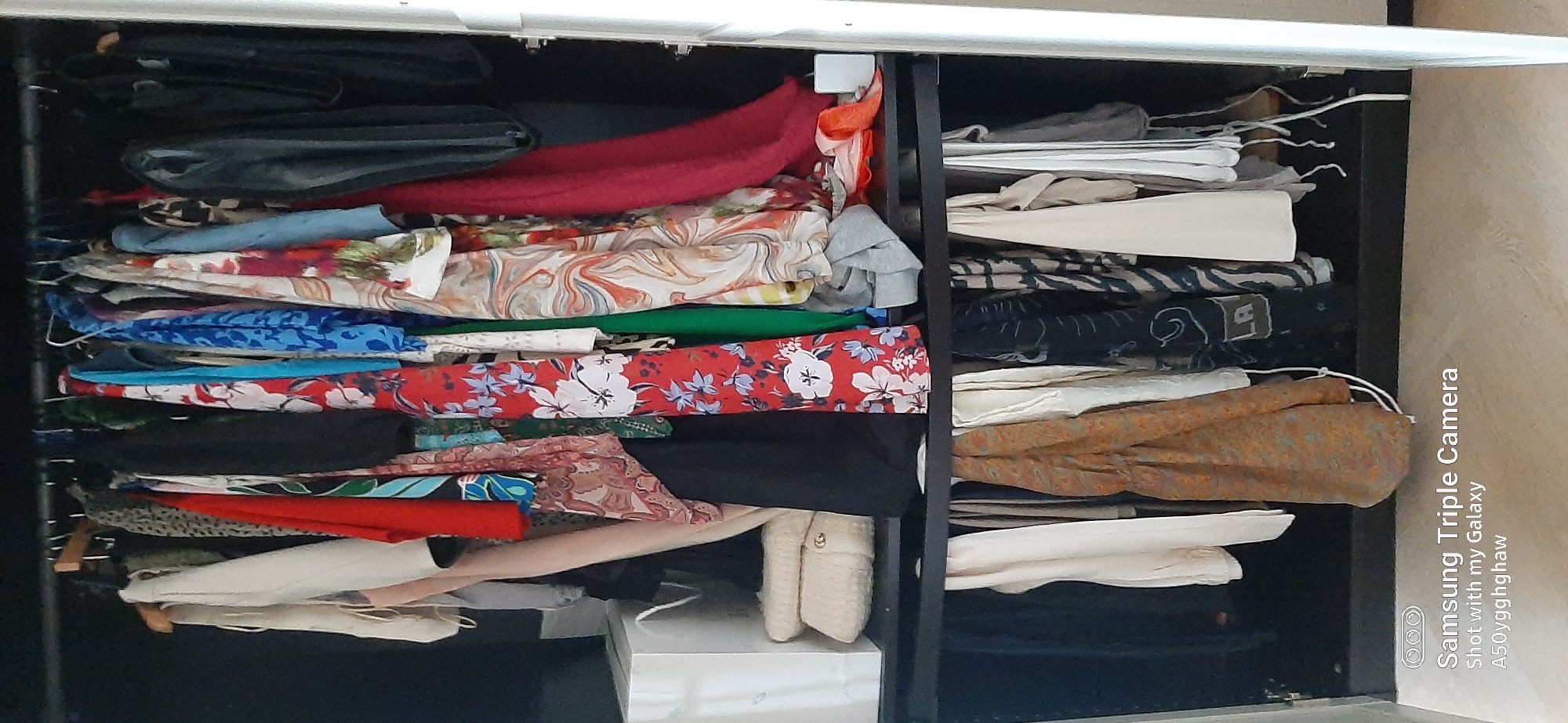 The image shows the interior of an open closet filled with various garments hanging on a rod. The closet is dark inside, with a shelf dividing the hanging space. On the shelf, to the left, is a white box and a stack of beige folded items. To the right of the shelf, there is a white object that appears to be an electrical outlet or a small device. The clothing items include dresses, skirts, and blouses in a variety of colors and patterns, such as floral prints, abstract designs, and solid colors like red, blue, green, and beige. Some of the garments are made of sheer material, while others appear to be thicker fabrics. Below the shelf, more clothes are hanging, including a brown patterned garment and several white and dark-colored items. The closet door frame is visible on the right side of the image. A portion of a beige floor is visible at the bottom of the image, with the text "Samsung Triple Camera" and "Shot with my Galaxy A50yggghaw" imprinted on it, indicating that the photo was taken with a Samsung Galaxy A50. The image does not contain any discernible information or context to suggest its location within the city of Dubai, United Arab Emirates.