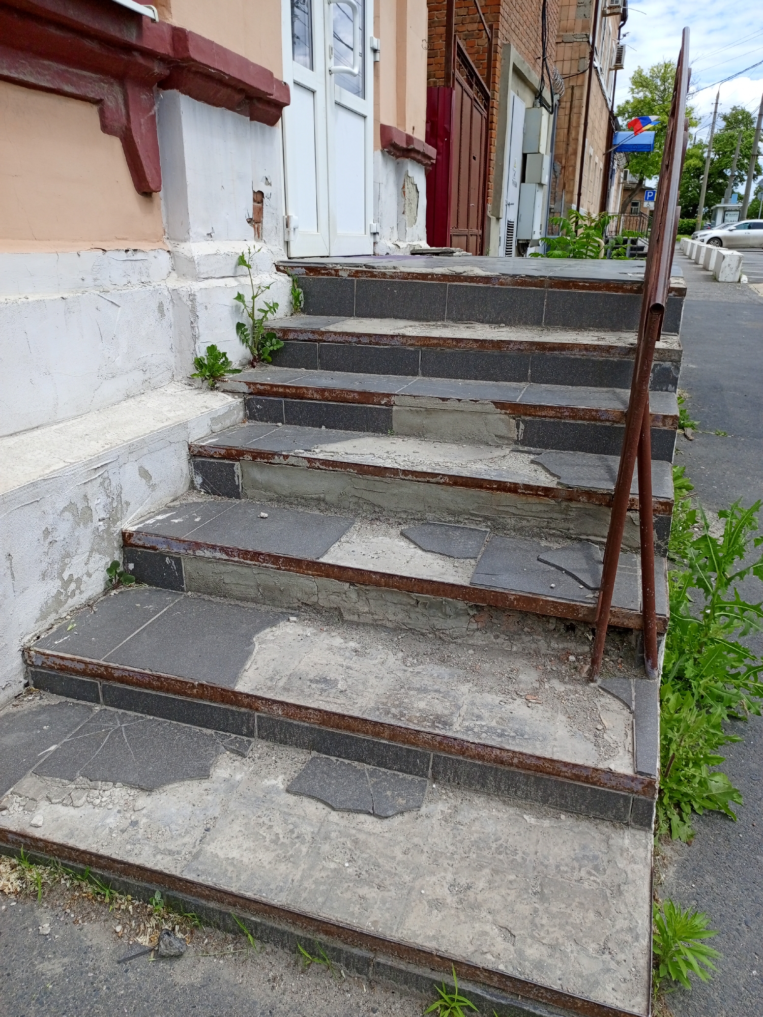 The image depicts a set of worn and damaged concrete stairs leading to an entrance of a building in an urban environment. The stairs are constructed with a combination of grey tiles and concrete, with metal edging. Several tiles are cracked or missing, revealing the underlying concrete. The steps are lined with weeds and other plants growing between the stairs and along the side. A rusted metal railing is positioned on the right side of the steps. The building adjacent to the stairs is made of brick, with a peach-colored facade on the left side. The scene is set outdoors, with a section of sidewalk visible in the lower right corner of the image. The sky is overcast, suggesting either early morning or late afternoon. A car is parked along the side of the street, beyond the stairs. A sign in blue with Ukrainian colors can be seen, along with a sign for parking. The building appears to be in a state of disrepair.