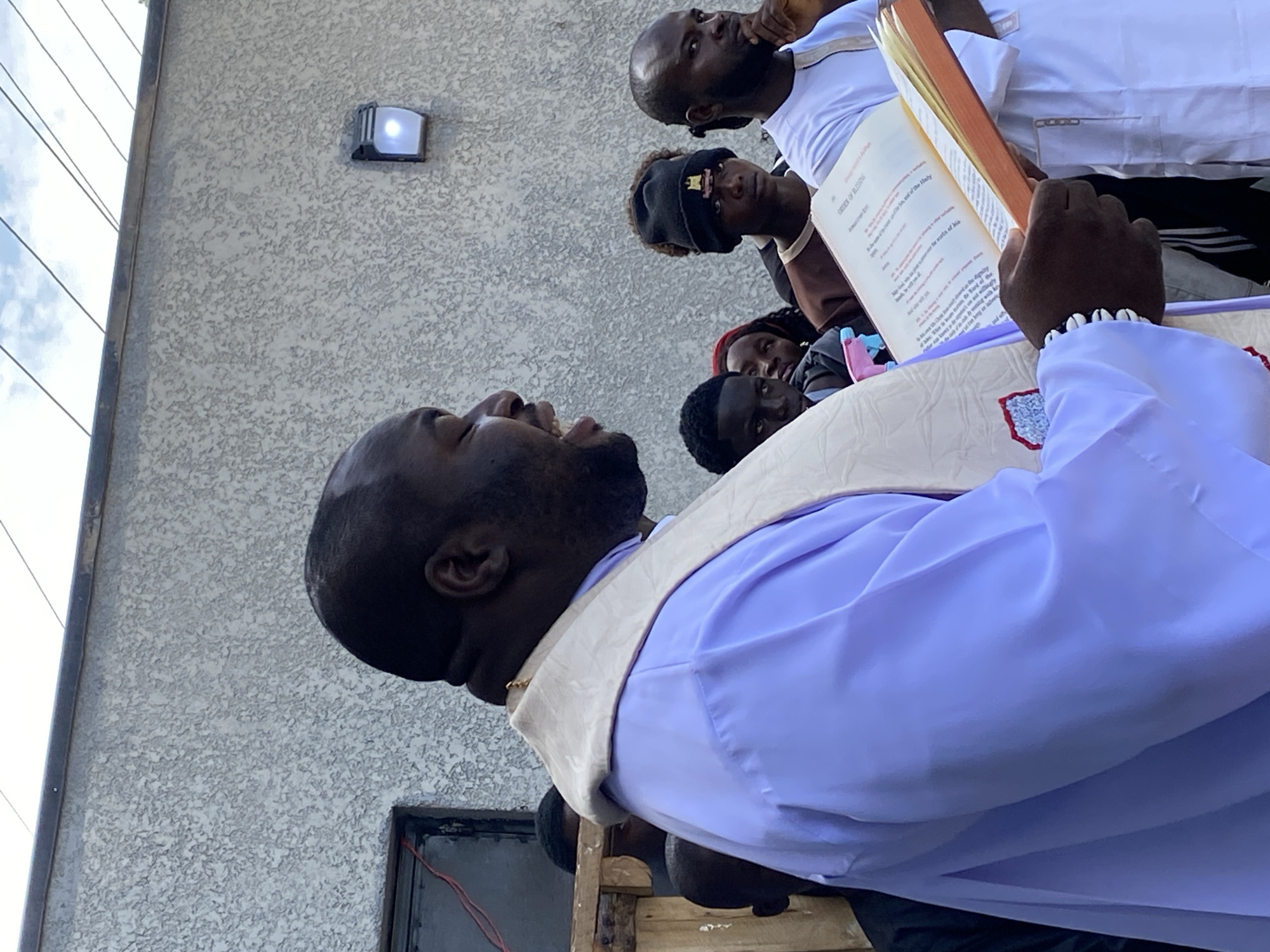A medium shot captures a man in a light purple robe and a cream-colored stole, reading from an open, orange-edged book. He is positioned in the foreground, to the left of the frame, with his mouth open as if speaking. Behind him, a group of individuals are gathered, their faces mostly visible. To the far right, another man in a white garment is also reading from a book. The background features a textured, light gray wall with a small, illuminated light fixture. The scene appears to be outdoors, possibly in an urban setting in Gura Top, Nigeria, with a glimpse of the sky and overhead wires visible at the top of the frame. The lighting suggests it is daytime.