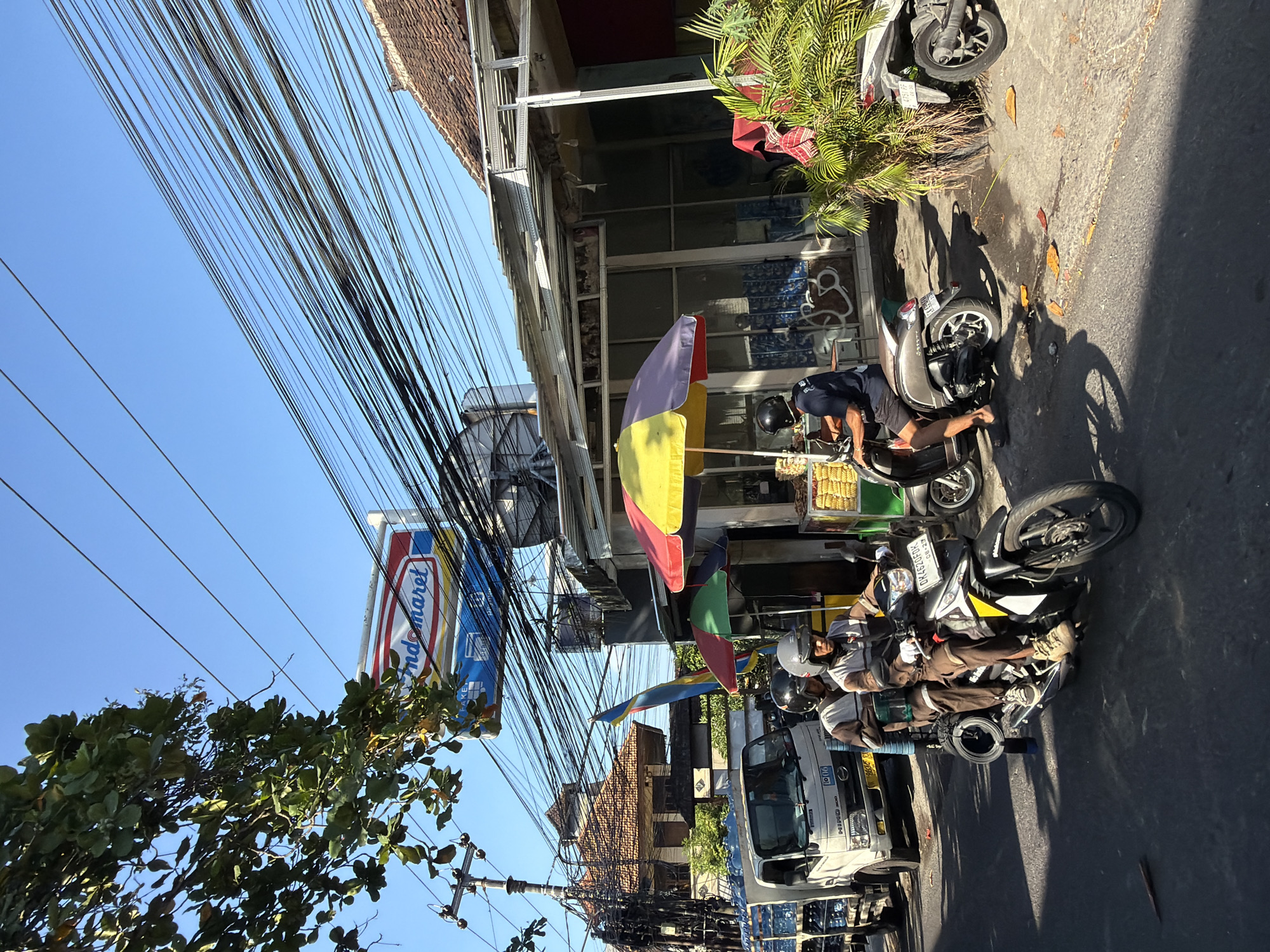 In North Kuta, Indonesia, a street scene unfolds under a bright blue sky, characterized by a dense network of overhead electrical wires. In the foreground, two individuals ride a dark-colored motorcycle. The rider in the front, wearing a grey helmet and a brown and white jacket, operates the vehicle. Behind him, a passenger with a black helmet and a similar jacket is seated. The motorcycle has a yellow and black design on its front fairing and displays a license plate that reads "DK 4620 FPK".
To the left of the motorcycle, a white truck laden with bottled water is parked on the road. The truck has "HINO 300" printed on its front. Further left, a utility pole with tangled wires and a few branches of a leafy tree are visible.
In the middle ground, a small food stall is set up under a colorful umbrella, featuring red, yellow, and green panels. The stall displays a rack of what appears to be fried snacks. Adjacent to the stall, a brown scooter is parked, with its rider wearing a dark blue shirt and black shorts, and a black helmet.
Behind the stall and the scooter, a building with a tiled roof and a partially constructed or damaged awning is visible. The building's facade features windows and some graffiti on the lower section. To the right, a lush green plant with palm-like leaves adds a touch of nature to the urban setting. The overall environment suggests a busy, possibly commercial street in a tropical region.