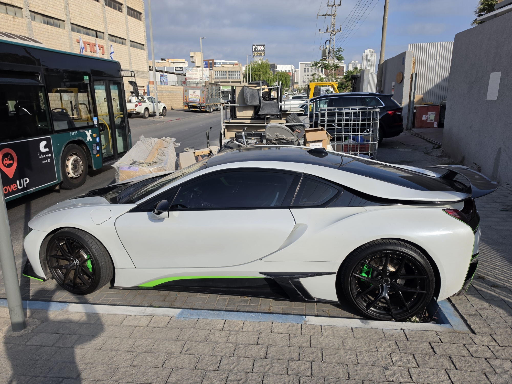 The image depicts a white and black BMW i8 parked on a paved area beside a street. The car has custom green accents and black wheels with green brake calipers. In the background, there is a bus with "MOVEUP" branding, a white pickup truck, and another truck loaded with various items, including chairs. A dark-colored SUV is partially visible behind the truck. The backdrop consists of buildings, including one with Hebrew signage and flags. Based on the presence of the "RIHON LEZION" logo on the bus, the location is Rishon LeZion, Israel. The sky is partially cloudy.