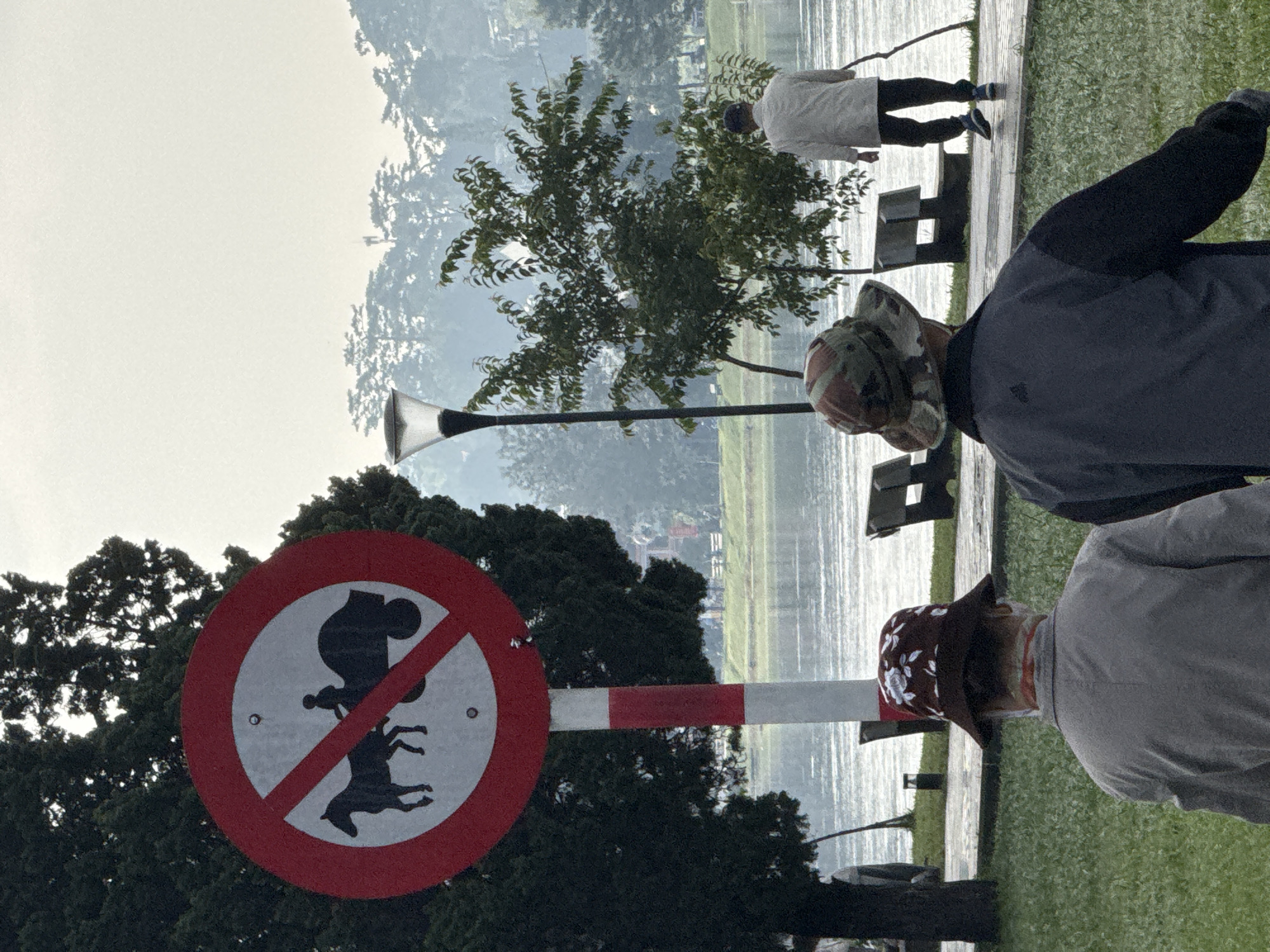 In Da Lat, Vietnam, a no horse-drawn carriage sign is prominently displayed. The sign is circular with a red border and a diagonal red line, indicating prohibition. Inside, a black silhouette depicts a horse pulling a carriage with a person in it. The sign is mounted on a red and white striped pole, positioned next to a lush green tree. In the foreground, two individuals are seen from behind, both wearing hats. One person wears a camouflage patterned hat and a dark jacket, while the other wears a brown floral hat and a light-colored jacket. Further in the background, a lake stretches out, reflecting the overcast sky. Several park benches are visible along the paved walkway by the lake. A man in a white shirt and dark pants is walking away from the viewer along the path, holding a walking stick. The overall atmosphere suggests a serene, outdoor setting, possibly a park or lakeside area.