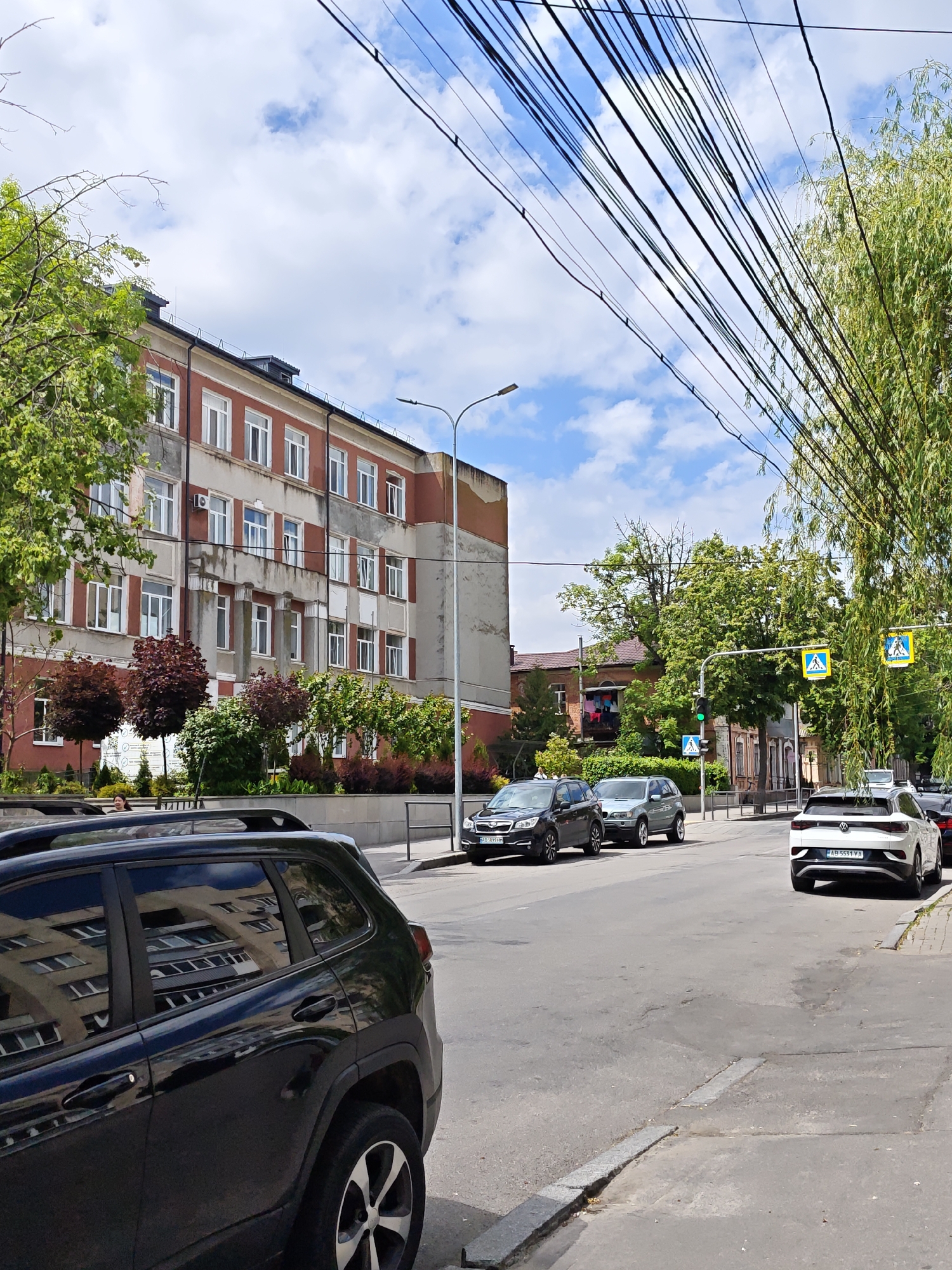 The image depicts a street scene in Vinnytsia, Ukraine, with several cars parked along the roadside. In the foreground, a black SUV is parked on the left side of the street. Across the street and further down, there's a silver SUV, a black SUV, and a white Volkswagen ID.4 with the license plate "AB 5531 YA." In the background, there is a tall, multi-story building with a red and beige facade. Pedestrian crossing signs are visible, hanging above the street. Electrical or communication wires stretch across the sky. There are various trees and greenery lining the street. The weather appears to be sunny with a partly cloudy sky.