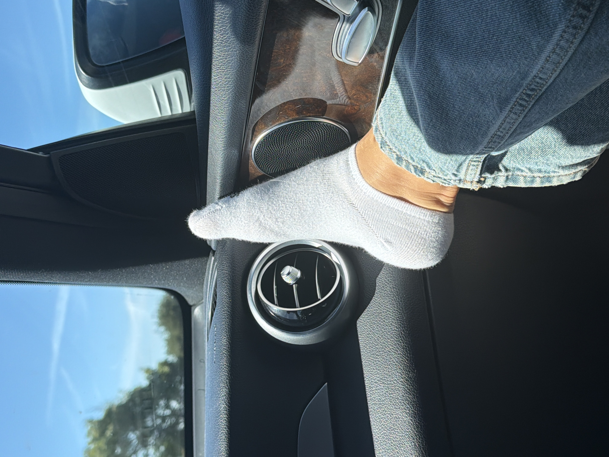 An individual's socked foot, clad in a white ankle sock and blue denim jeans, rests against the interior of a vehicle. The foot is positioned on the door panel, near a circular air vent with chrome accents and a textured speaker grille. The interior also features a section of wood-grain trim and a chrome handle. Outside the vehicle, visible through the window, is a clear blue sky with wispy clouds and blurred green trees. The scene suggests a sunny day, likely in a suburban or urban setting, but specific location in Oakland, United States of America cannot be determined from the image alone.