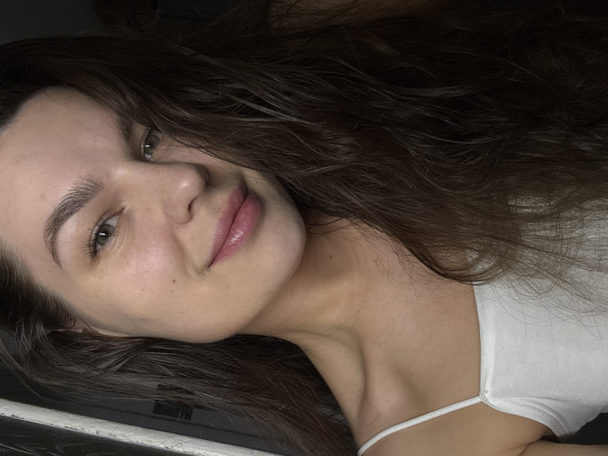 The image is a close-up, head-and-shoulders selfie of a young woman with long, dark, wavy hair. She is wearing a white tank top. Her skin is fair with some subtle freckles. Her eyes are green, and she has neatly shaped eyebrows. Her lips are full and appear to be lightly glossed. The background is dark and indistinct, with a vertical element on the left that could be a doorway or frame. There is no indication of the city of Chaikovsky, Russia, in this image.