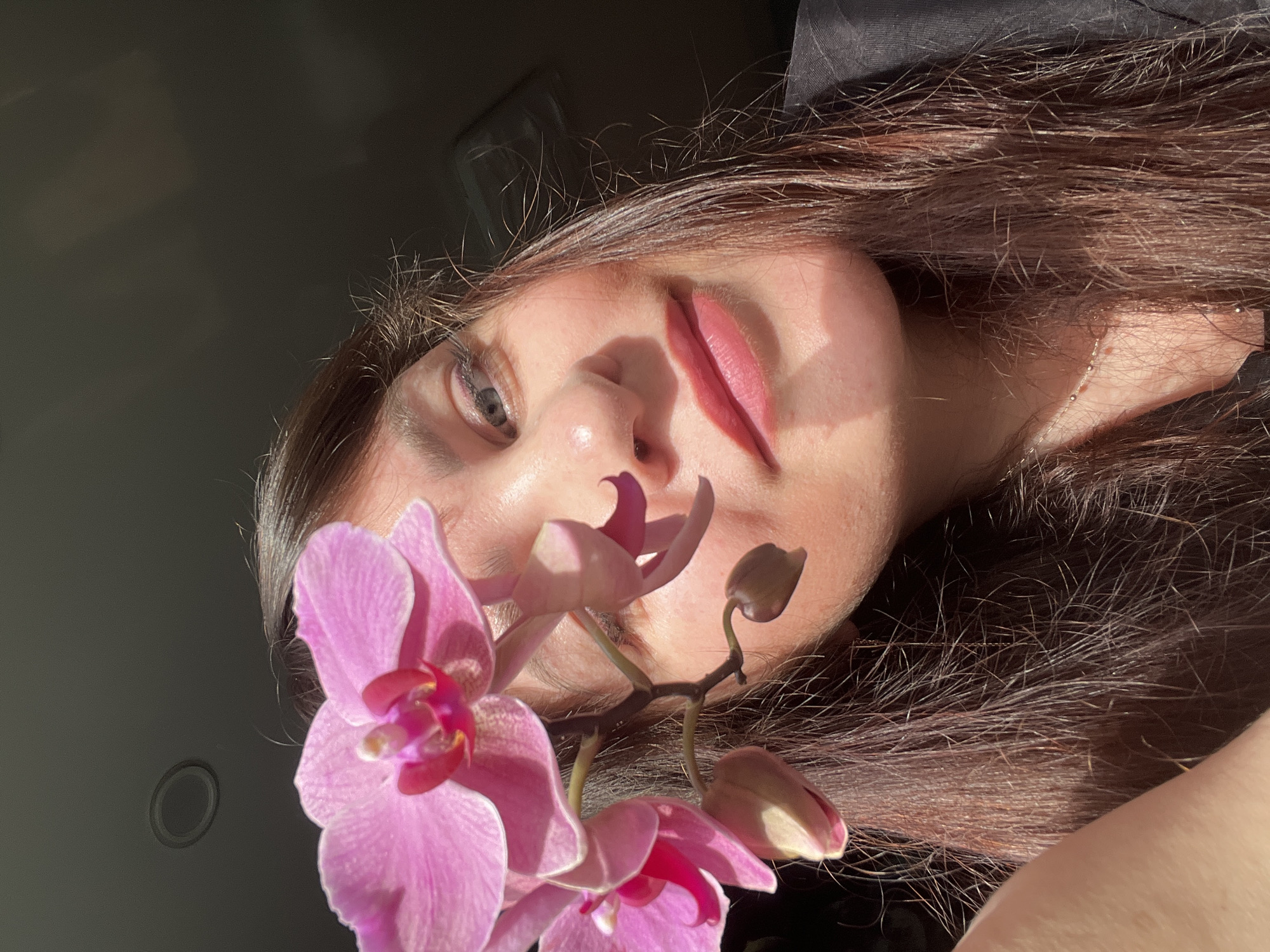A close-up, high-angle portrait captures a person, likely female, with long, dark brown hair, illuminated by strong, direct sunlight. Her face, angled sideways and seen from the nose up, features her left eye looking towards the viewer and her lips slightly parted. Prominent shadows are cast across her face by the bright light, which also highlights individual strands of her hair.
In the foreground, a branch of vibrant pink Phalaenopsis orchids is positioned, displaying several open blooms and buds. The orchids partially obscure the lower part of the person's face, creating an artistic composition. The background is a dark, uniform surface, possibly the interior of a vehicle or a dimly lit room, providing a stark contrast to the brightly lit subject and flowers.
The direct sunlight suggests a sunny day, likely morning or late afternoon. The overall mood is serene and aesthetic, emphasizing natural light, floral beauty, and human features. No text is visible in the image. This scene was captured in Noviy, Russia.