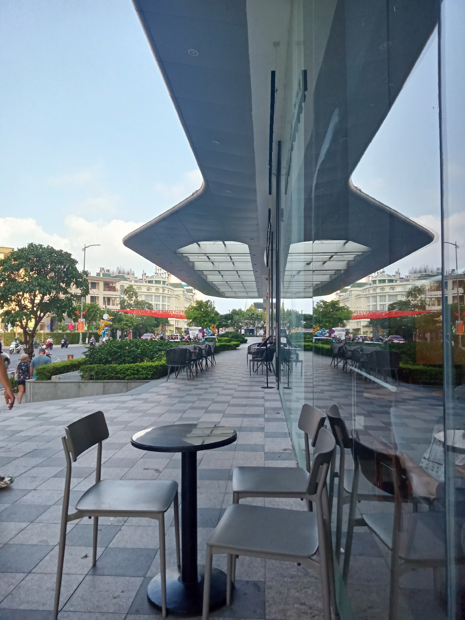 An eye-level shot captures an outdoor cafe or restaurant seating area in Nha Trang, Vietnam, during what appears to be late afternoon or early evening. The foreground features a small, round dark table and two modern chairs with light grey seats and backs, positioned on a paved area composed of alternating grey and black square tiles. A large, distinctive architectural canopy with recessed lights extends overhead.
The building's expansive glass facade dominates the right side of the image, creating prominent reflections. These reflections mirror the bustling street scene opposite, showing a line of tables and chairs, lush green shrubbery, and the distant street. The actual street, visible on the left side, shows people walking, including a person in a blue shirt and another in a floral patterned outfit. Numerous motorbikes and a few cars traverse the road, and a cluster of red festive decorations, possibly lanterns or banners, are strung across the street further down. Residential or commercial buildings with light-colored facades and green rooftops line the street beyond the greenery.
The strong reflections on the glass facade create an almost surreal, doubled image of the surrounding environment, including the overhead canopy which appears inverted in the reflection. The sky is bright blue with scattered white clouds, suggesting clear weather. The overall atmosphere is that of a lively urban setting, with daily activities taking place. No discernible text is visible.