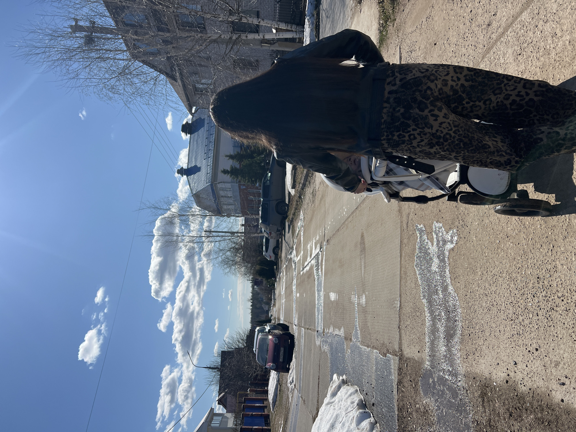 A person with long, dark hair and wearing a dark jacket and leopard-print pants is pushing a white stroller along a sidewalk on a sunny day. Patches of snow are visible on the side of the road and sidewalk. The street is wet, reflecting the sunlight. Several cars are parked along the side of the street. Buildings are visible in the background. The location is Noviy, Russia.