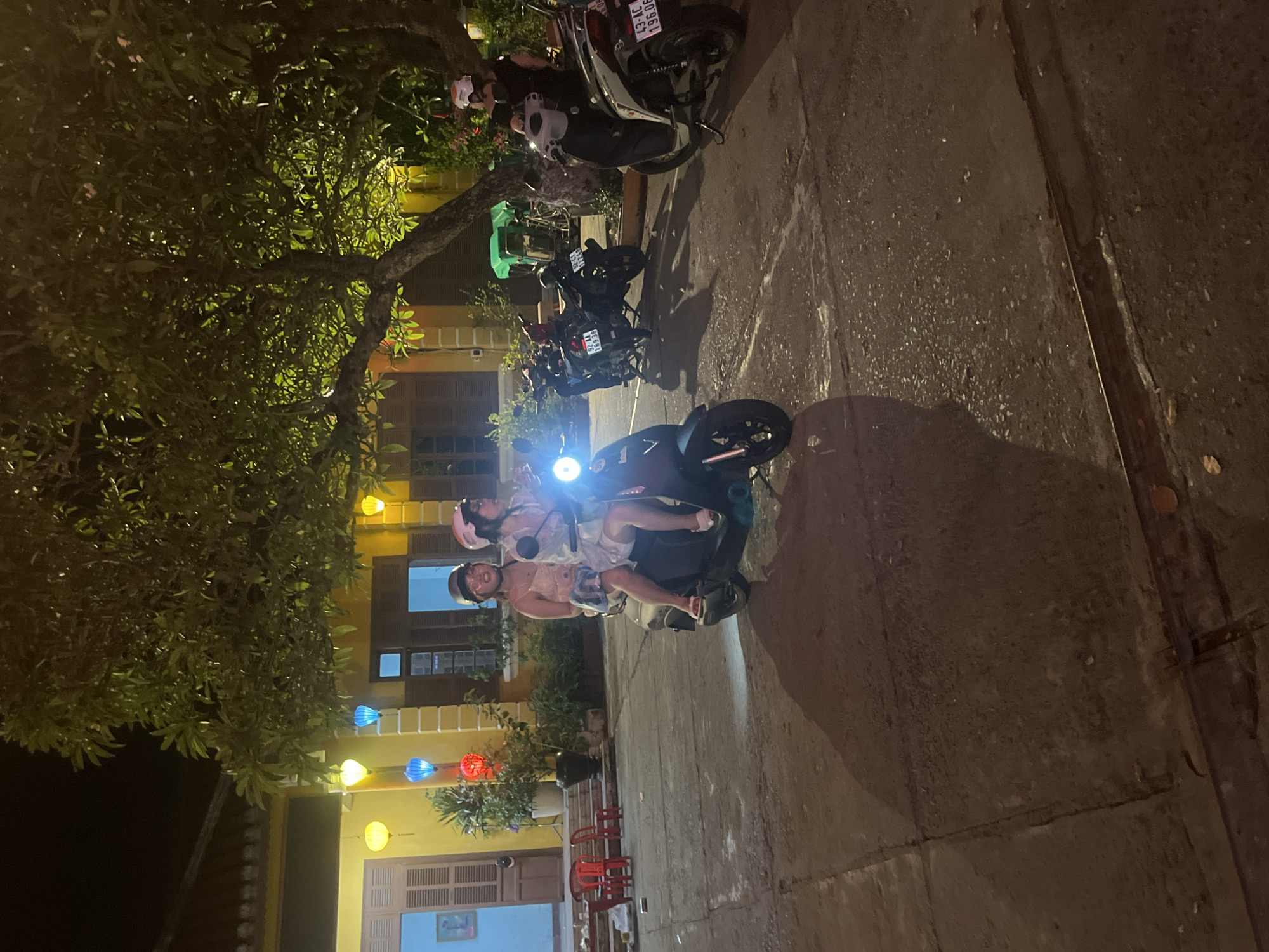 Two young women, wearing helmets, ride on a dark-colored scooter at night in what appears to be a street in Hội An Ward, Vietnam. The woman in front is wearing a white dress and the woman behind her is wearing a pink patterned dress. They are both looking forward, with the rider steering. The scooter's headlight is illuminated. In the background, a yellow building with closed shutters and balconies is visible, decorated with colorful lanterns of red, yellow, and blue. A large tree with green foliage extends over the scene from the right. Other scooters are parked nearby. The ground is paved with concrete. The lighting suggests it is nighttime.