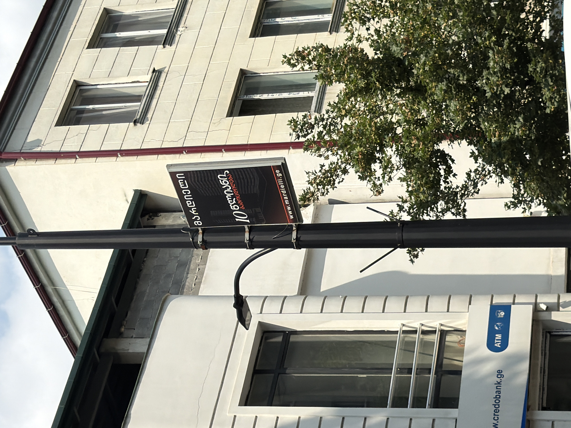 This is an eye-level, full outdoor shot of a street in Tbilisi, Georgia, on a partly cloudy day. In the foreground, a black lamppost stands upright and is adorned with a sign. The sign is black with white text in Georgian and English, featuring the URL "www.mardielli.ge". To the left of the lamppost, a white building with a blue sign that reads "credobank.ge ATM" is visible. This building has a large window with a metal railing. The background features a multi-story building with white walls and numerous windows. A lush green tree with many leaves partially obstructs the view of the background. The overall scene depicts a typical urban streetscape with commercial elements.