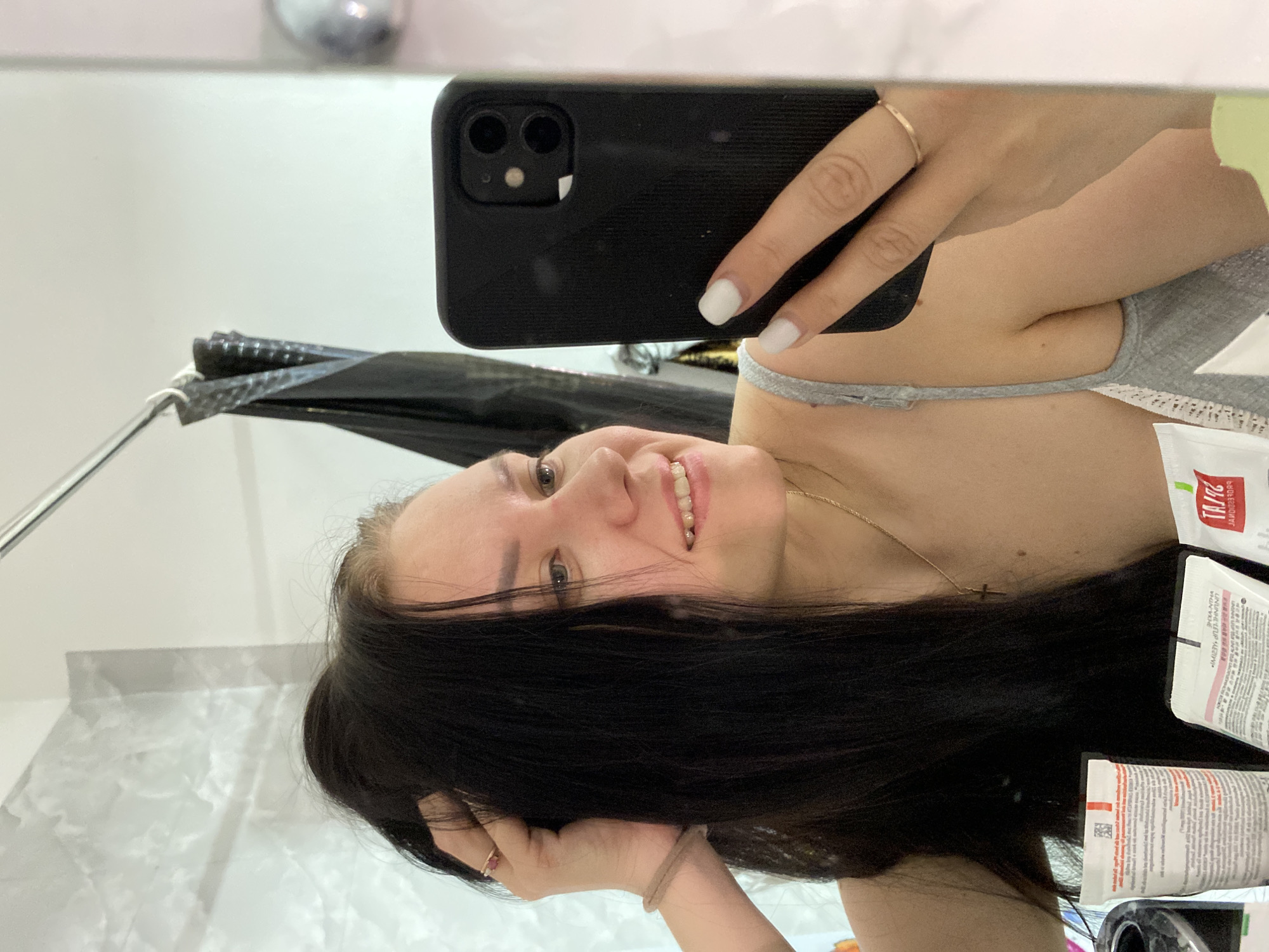 A woman is taking a selfie in a bathroom mirror. She is holding a black smartphone in her right hand. The woman has long, dark hair and is wearing a gray tank top. A cross necklace is visible. Several cosmetic tubes and bottles are placed on the bathroom counter near her. The reflection in the mirror includes a towel rack with a black towel. The bathroom has light-colored walls and a marble countertop. It's not possible to confirm the location is Chaikovsky, Russia, from the image alone.