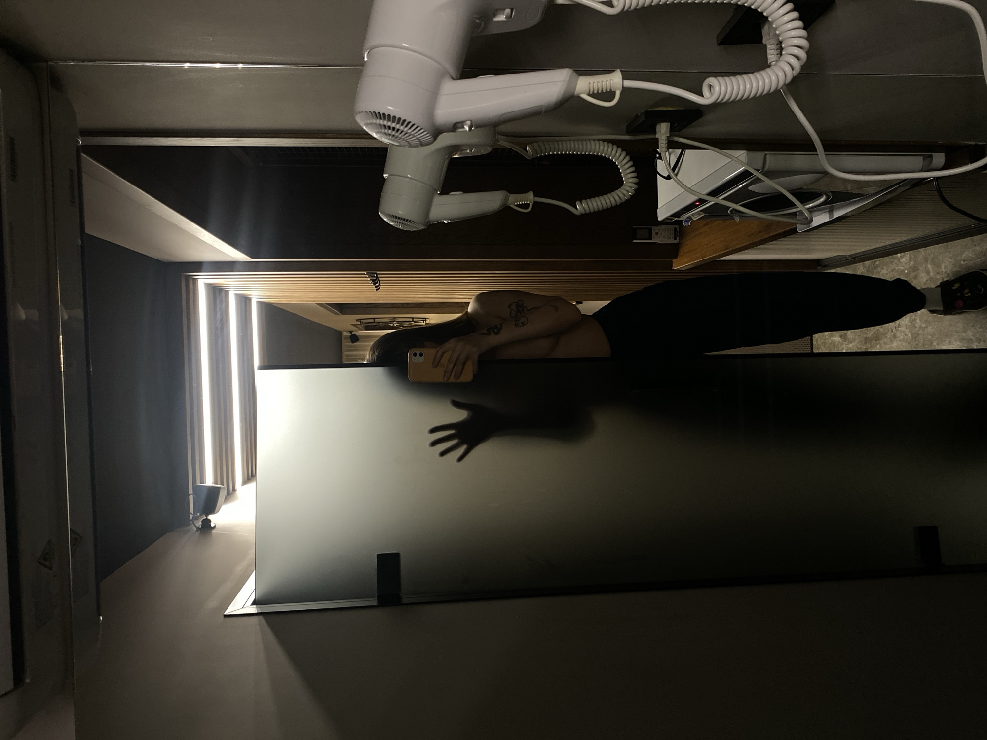A person is reflected in a bathroom mirror. The person has a tattoo on their arm and is holding a smartphone. The mirror is located in a bathroom, as evidenced by the presence of a hair dryer and straightener mounted above the mirror. The image appears to be taken indoors, with a glimpse of an interior space illuminated by vertical lights in the background. There is no information to suggest the location is in Votkinsk, Russia.