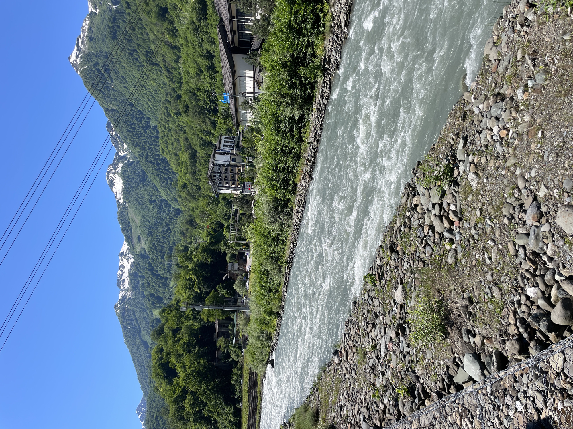 The image shows a wide, fast-flowing river in Estosadok, Russia. The river is bordered by rocky banks on the left and a line of lush green trees and vegetation on the right. In the background, several multi-story buildings, some with dark roofs and white facades, are visible nestled amongst the trees. Beyond the buildings, majestic mountains rise up, their peaks capped with patches of snow against a clear blue sky. Power lines stretch across the upper portion of the image. The scene suggests a natural landscape integrated with human development, likely a resort area. There are no people visible in the image.