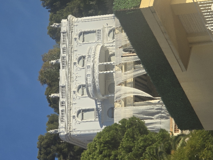 A white, ornate building with arched windows and balconies stands against a blue sky and trees. The building is partially obscured by greenery at the bottom and a green, artificial covering on the right, which is connected to a yellow structure with a railing. The architectural style appears to be classical or baroque, with intricate details and decorations. The composition is an upward-looking shot, highlighting the building's façade.